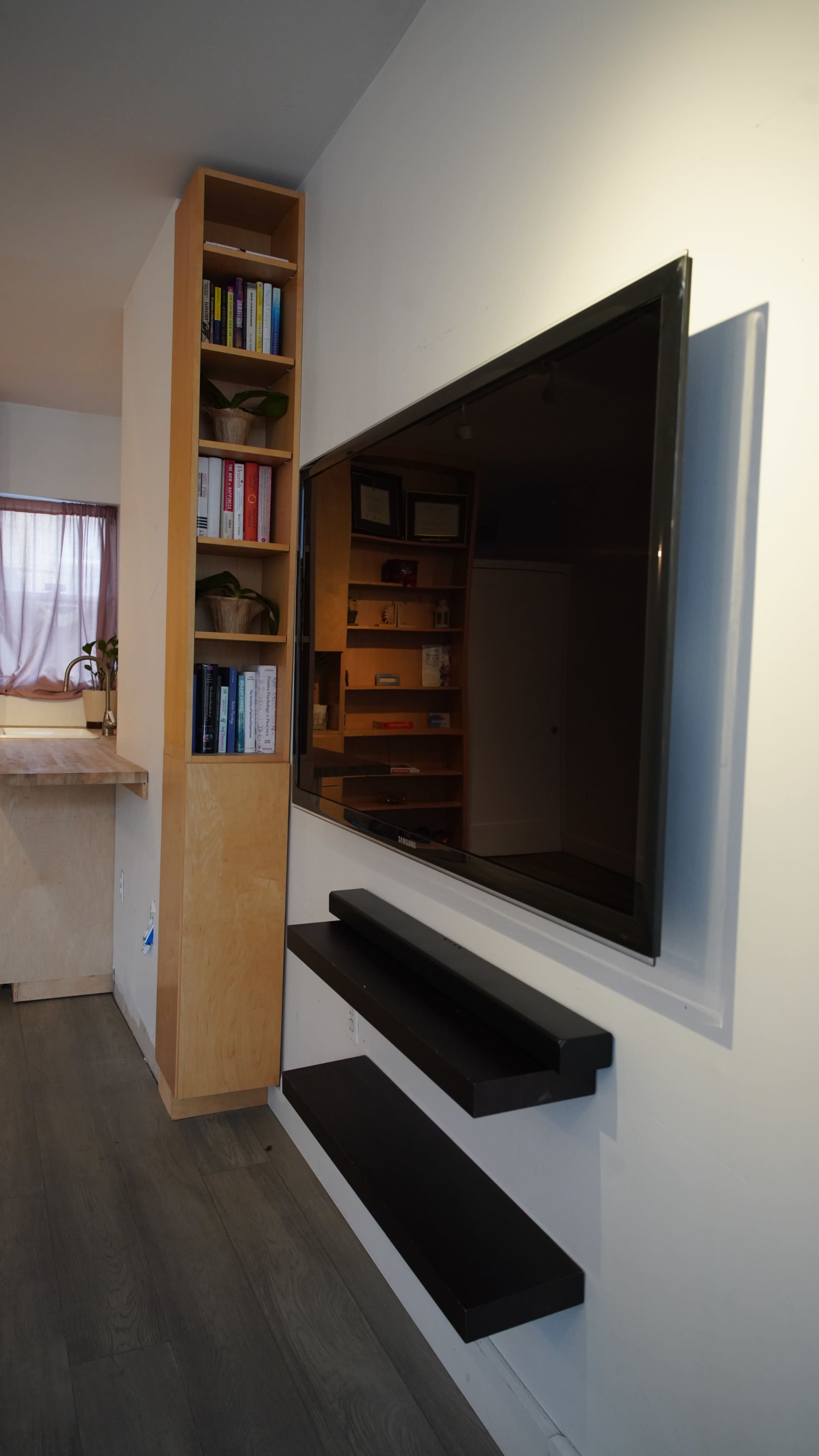 The image shows a modern living space featuring a mounted flat-screen television on a wall, with a narrow shelf below it, and a wooden bookshelf adjacent to it.