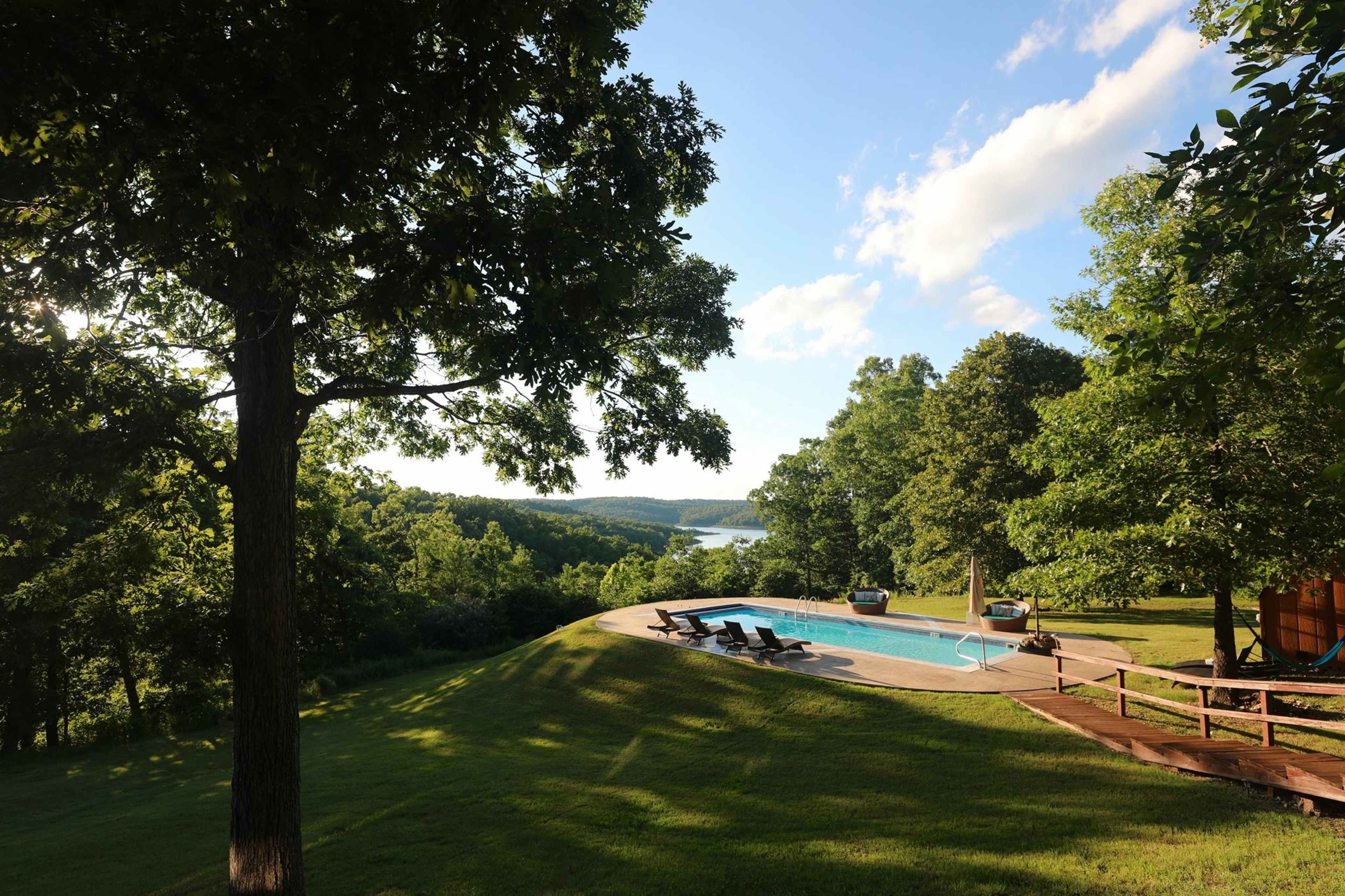 A swimming pool is situated on a lawn overlooking a valley with trees and a body of water in the distance.