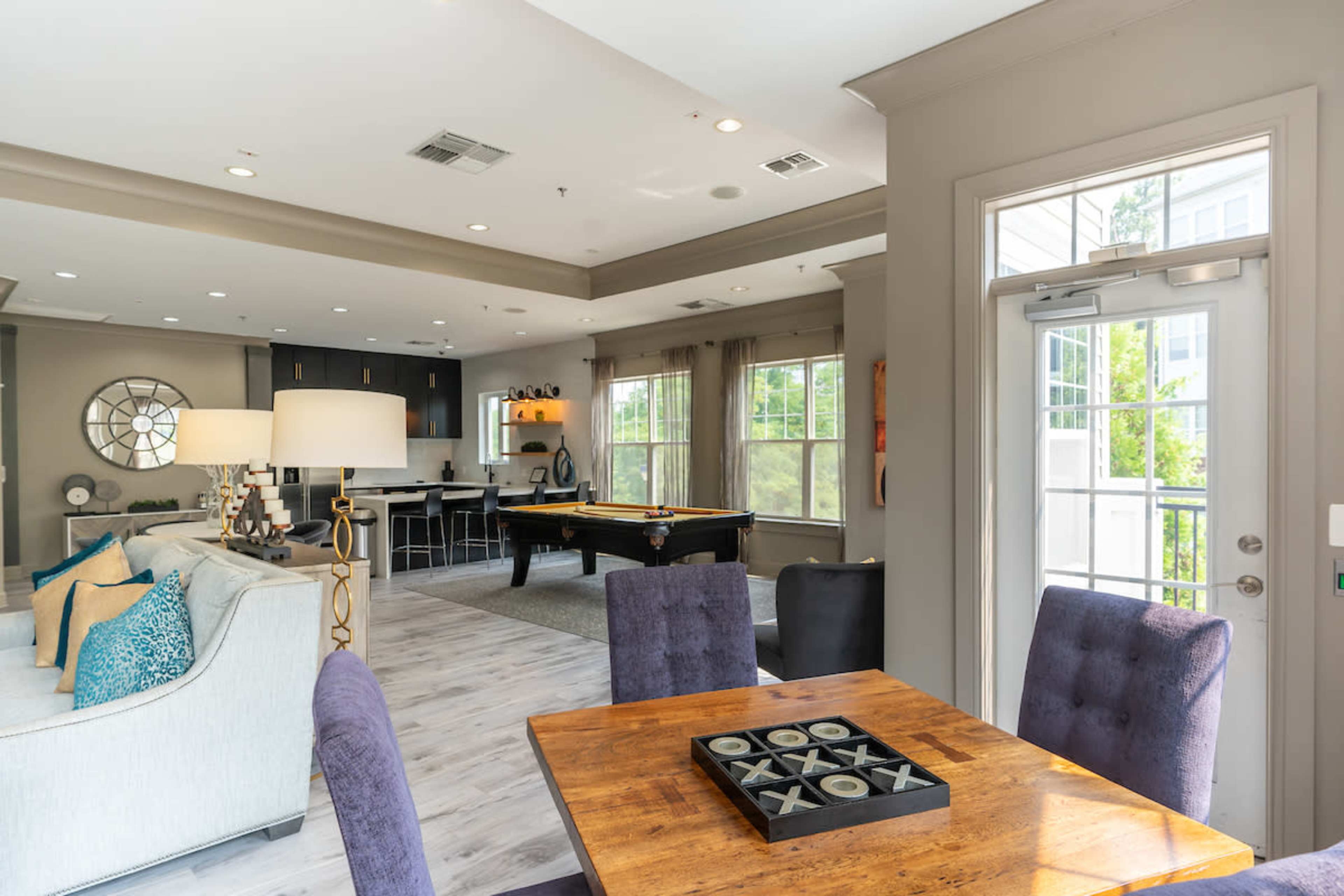 The image shows a modern living area featuring a dining table with chairs, a pool table, and an open kitchen area with large windows.