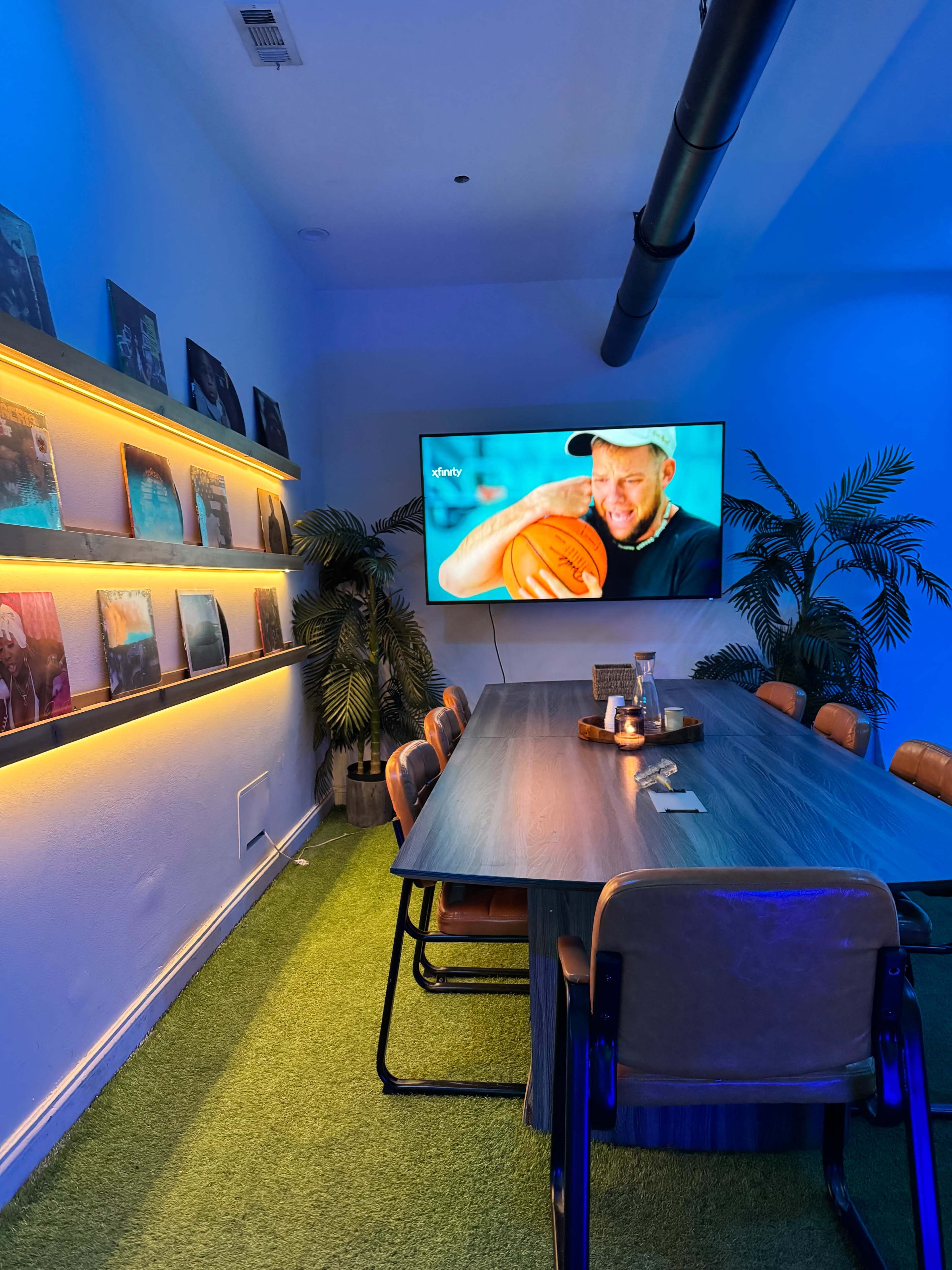 A conference room features a long wooden table surrounded by chairs, with a TV displaying a man holding a basketball and illuminated by blue lighting and palm plants along the walls.