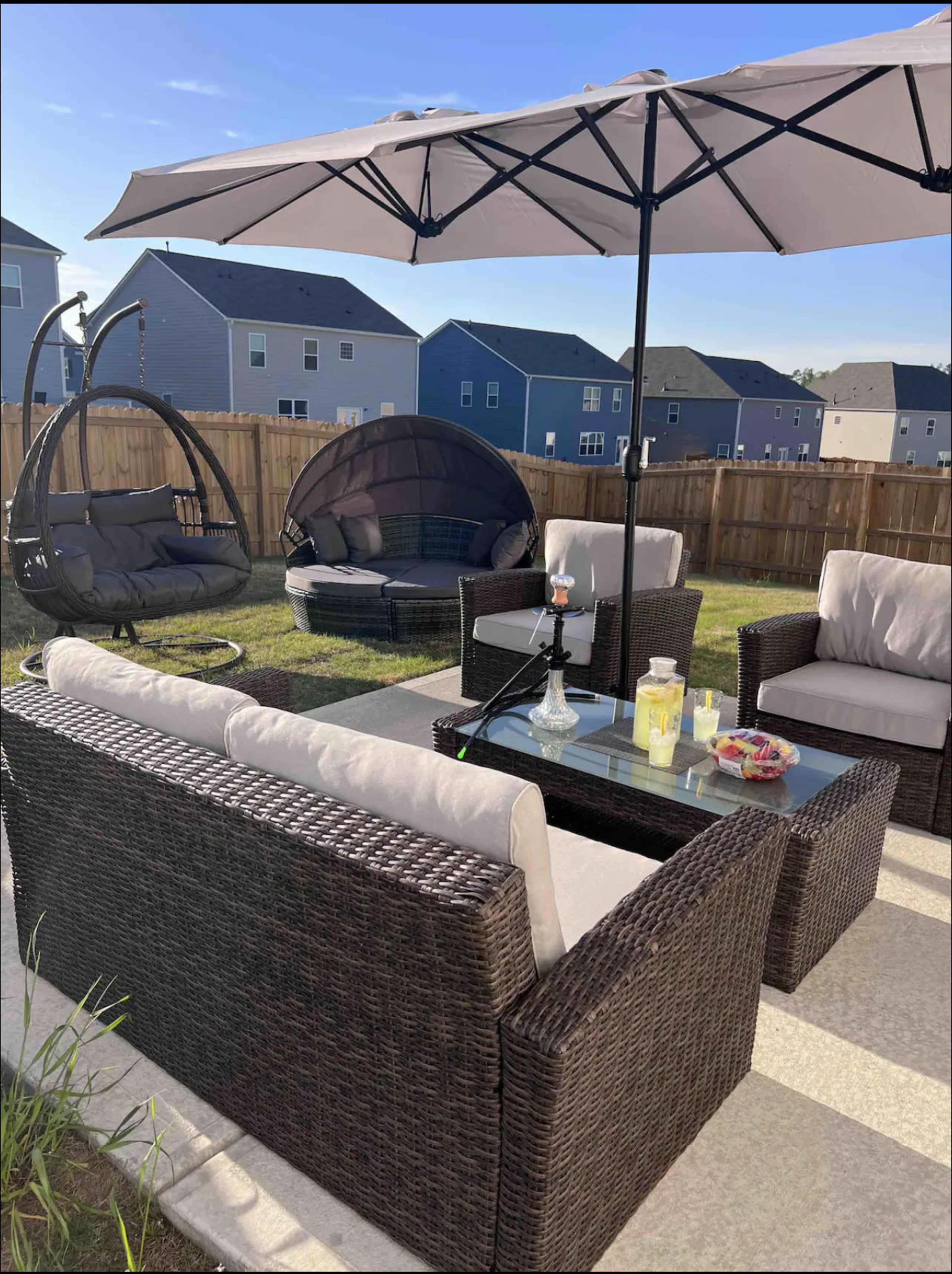 An outdoor patio area with a seating arrangement that includes wicker furniture, a glass coffee table, and a large umbrella, set against a wooden fence and several residential homes in the background.