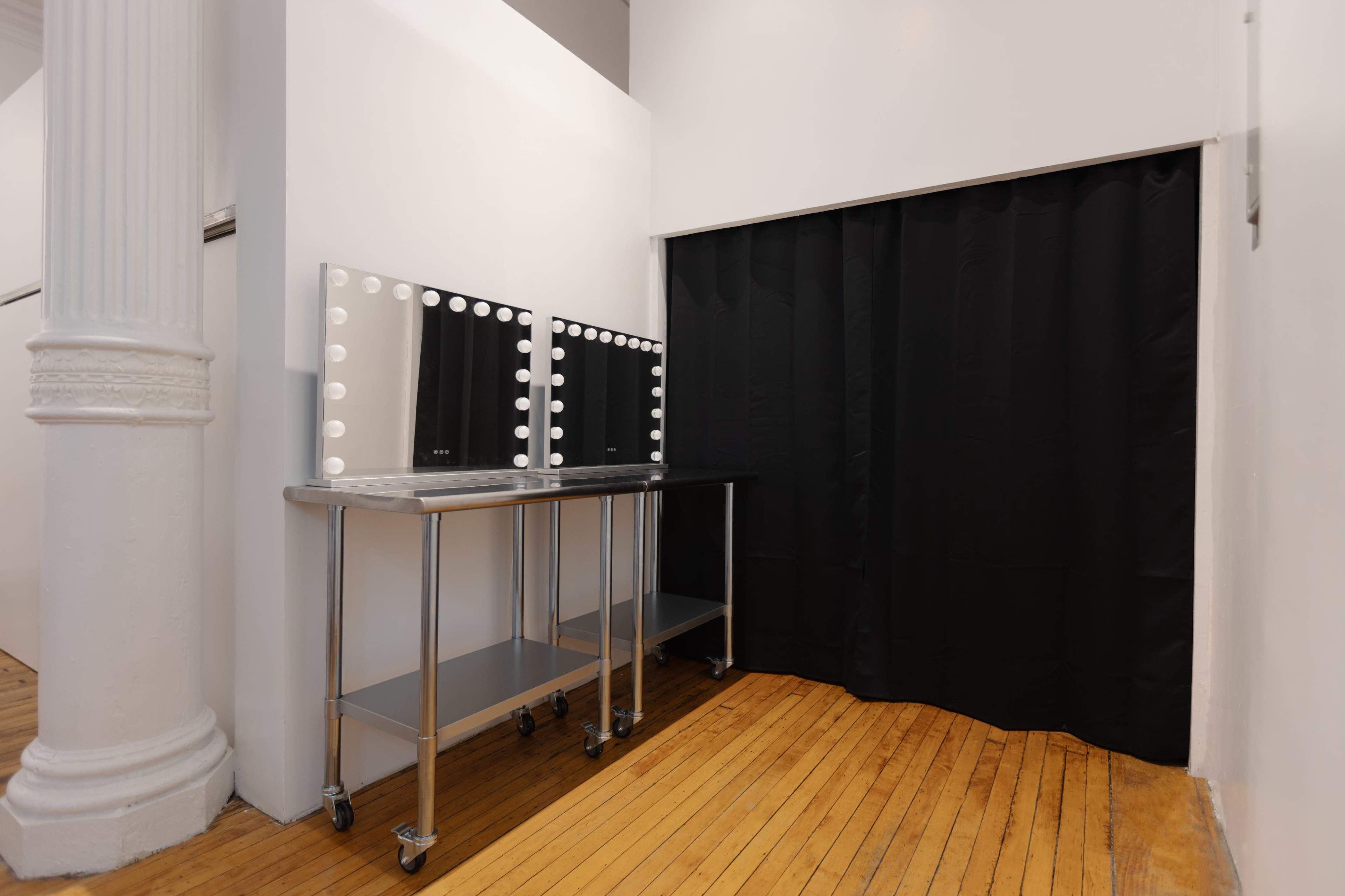 The image shows a room with two lit mirrors on a metal table beside a black curtain.