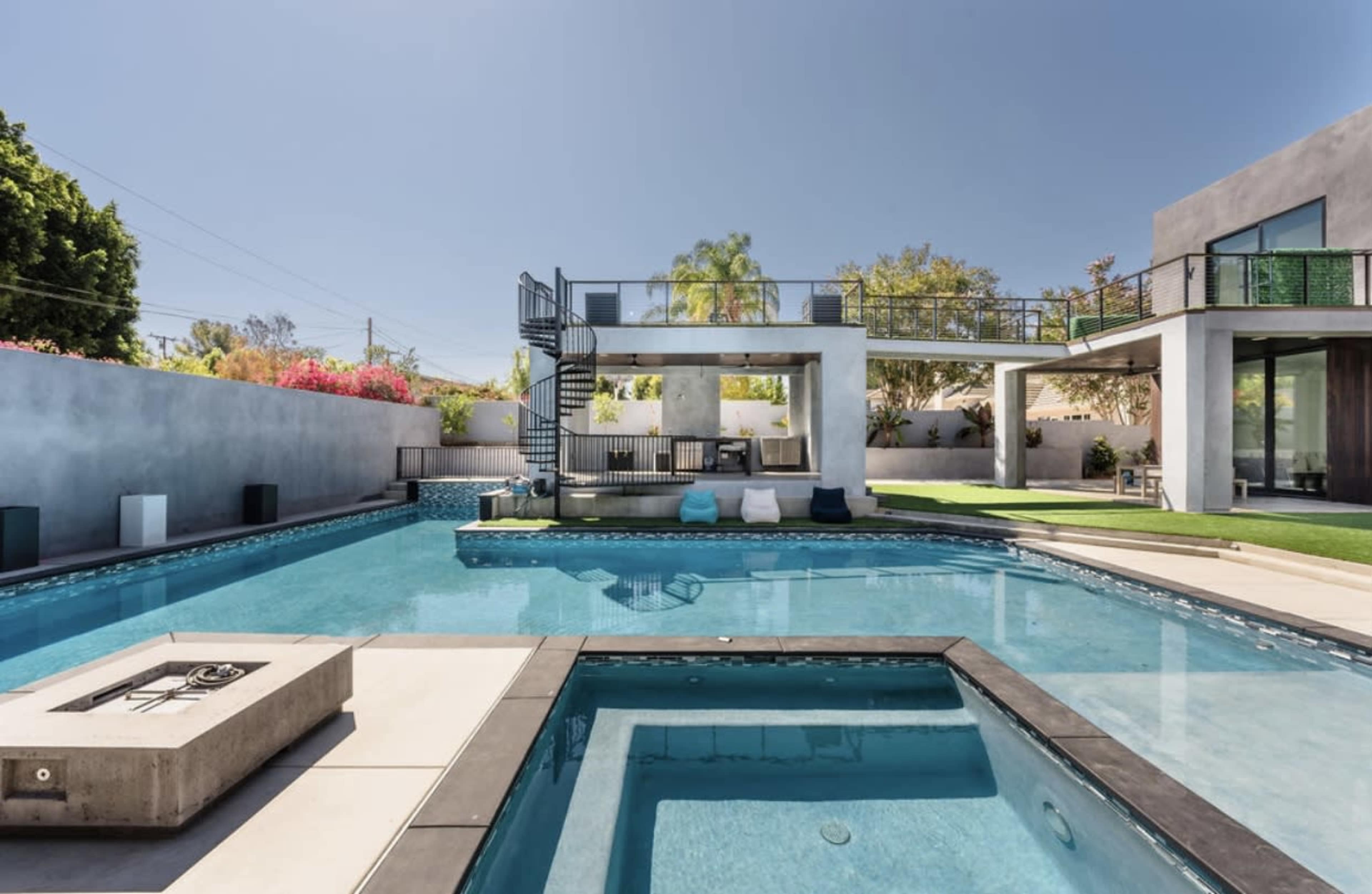 A modern home with an expansive swimming pool features a hot tub, landscaped areas, and multiple outdoor lounging spaces.