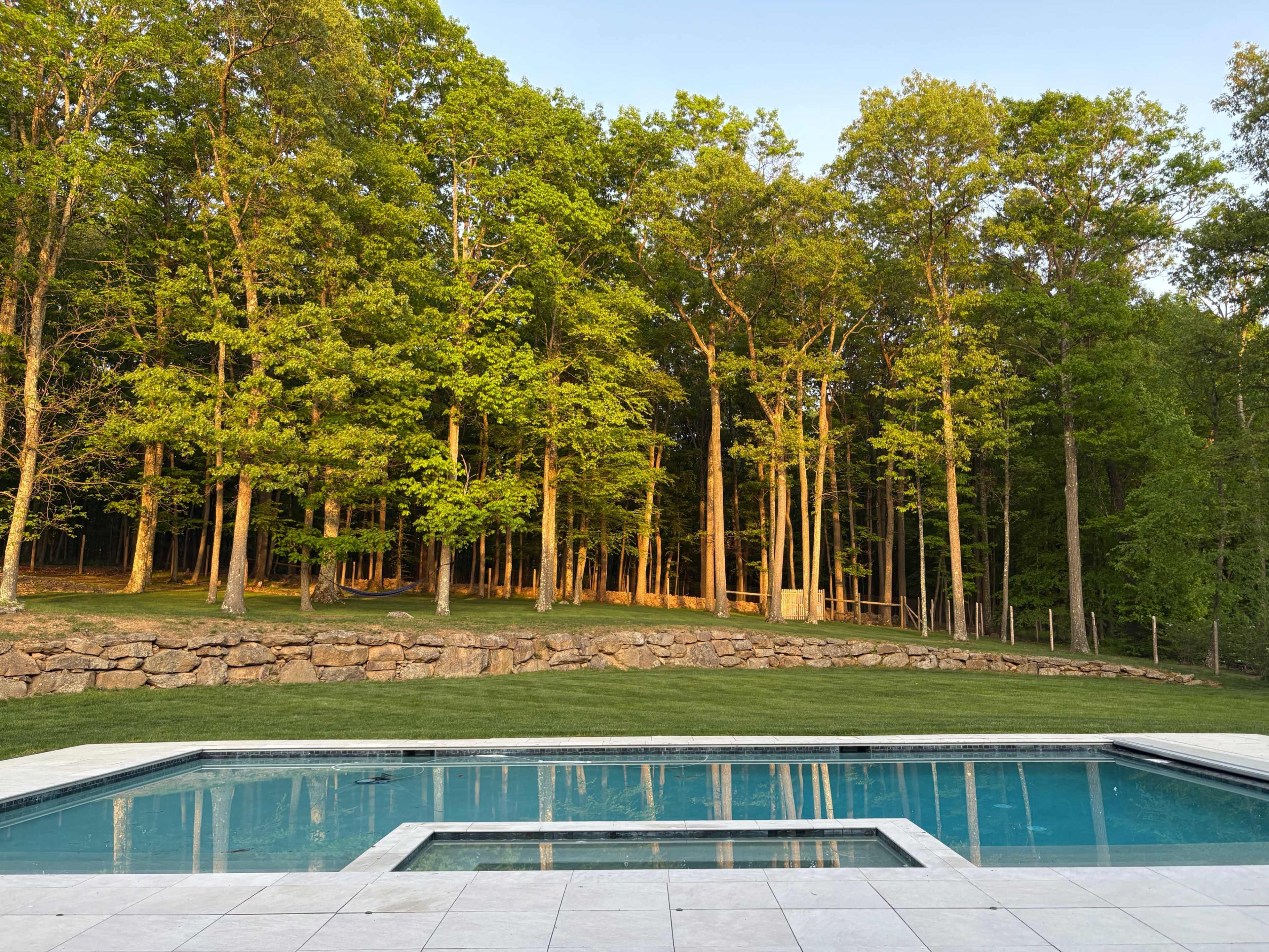 A rectangular swimming pool is situated in front of a stone wall, with a backdrop of sunlit trees in a forested area.