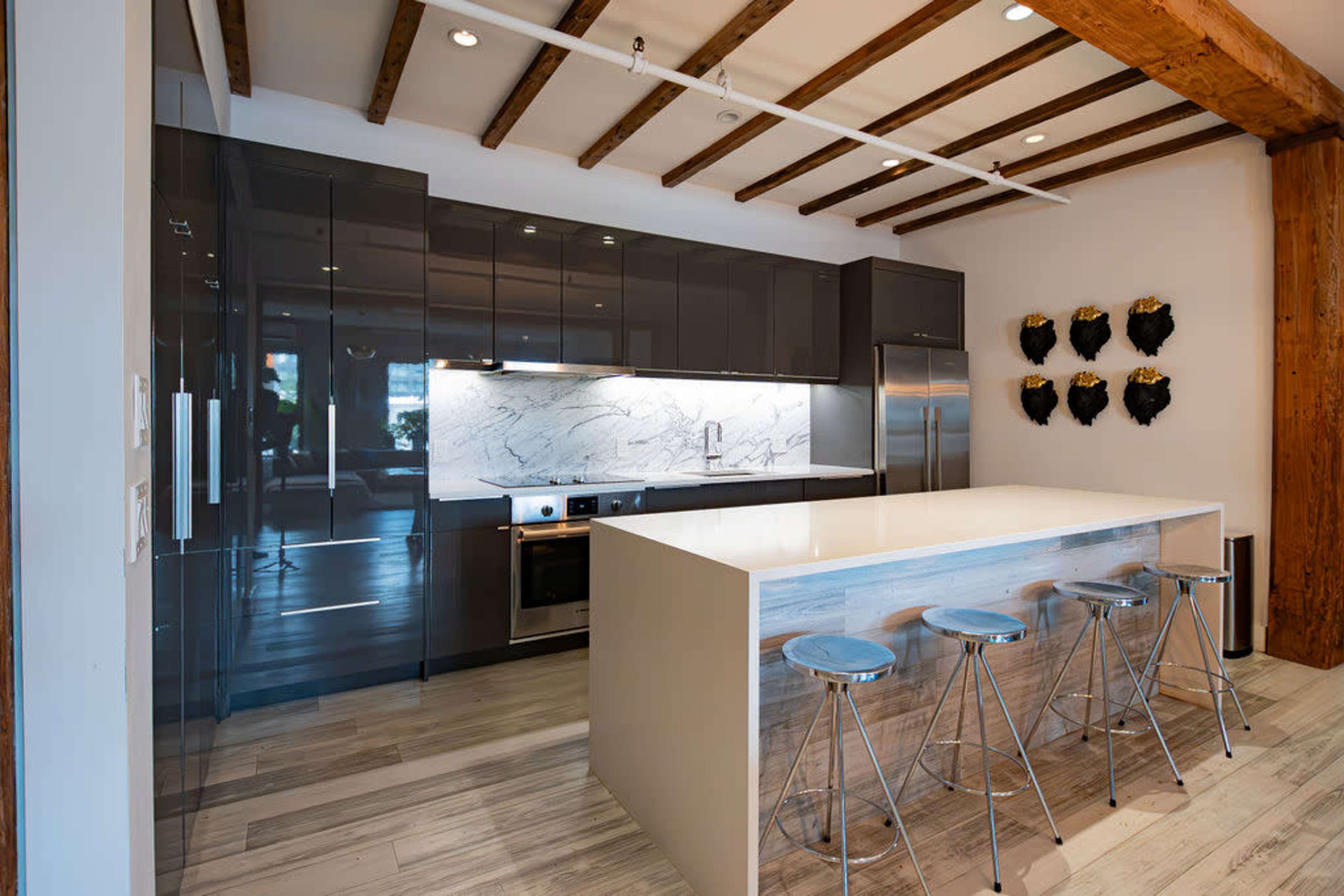 The image shows a modern kitchen with dark cabinetry, a marble backsplash, stainless steel appliances, and a central island with four metal bar stools.