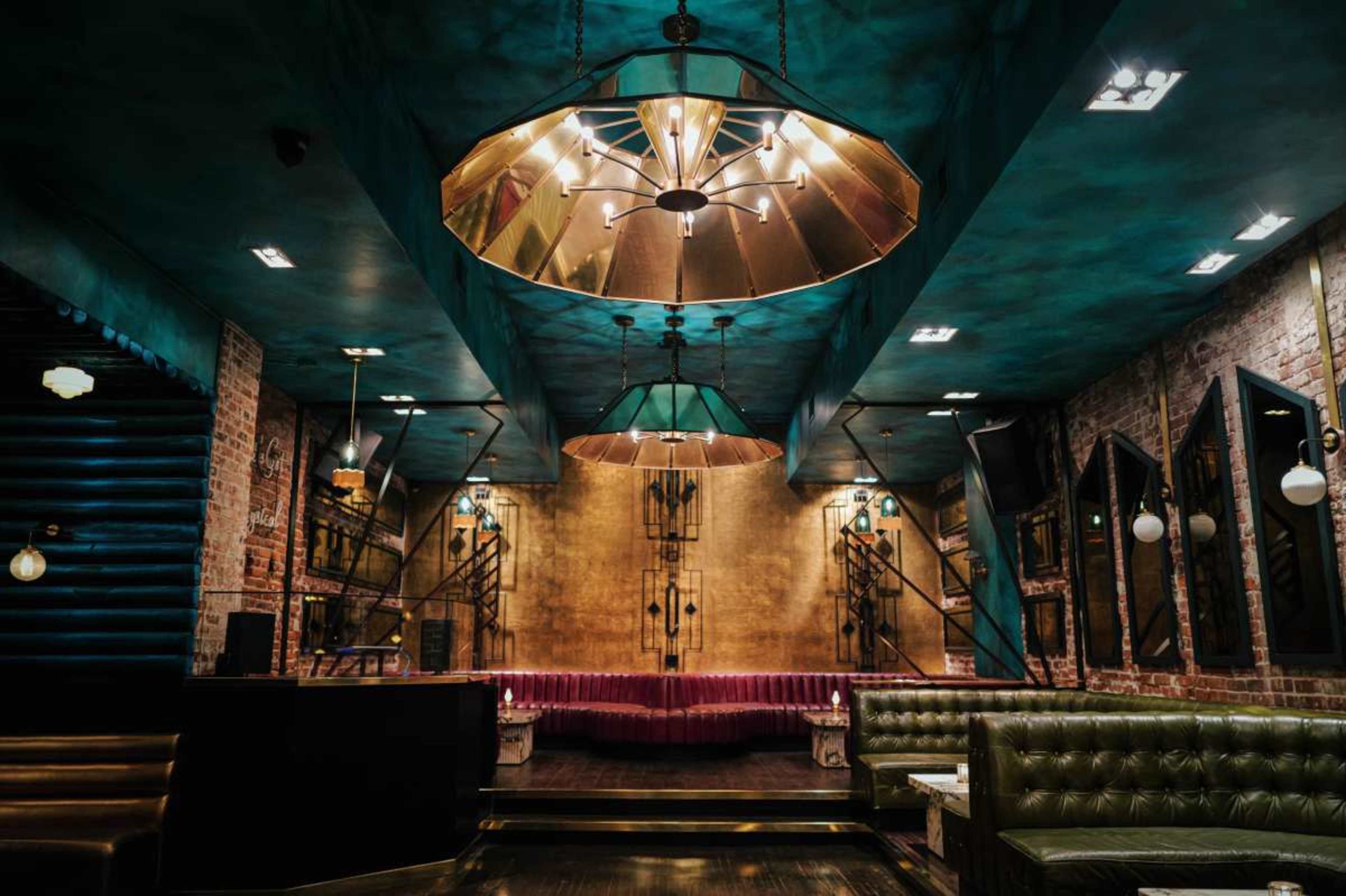 The image shows a stylish restaurant interior featuring green leather seating, exposed brick walls, and large decorative light fixtures.