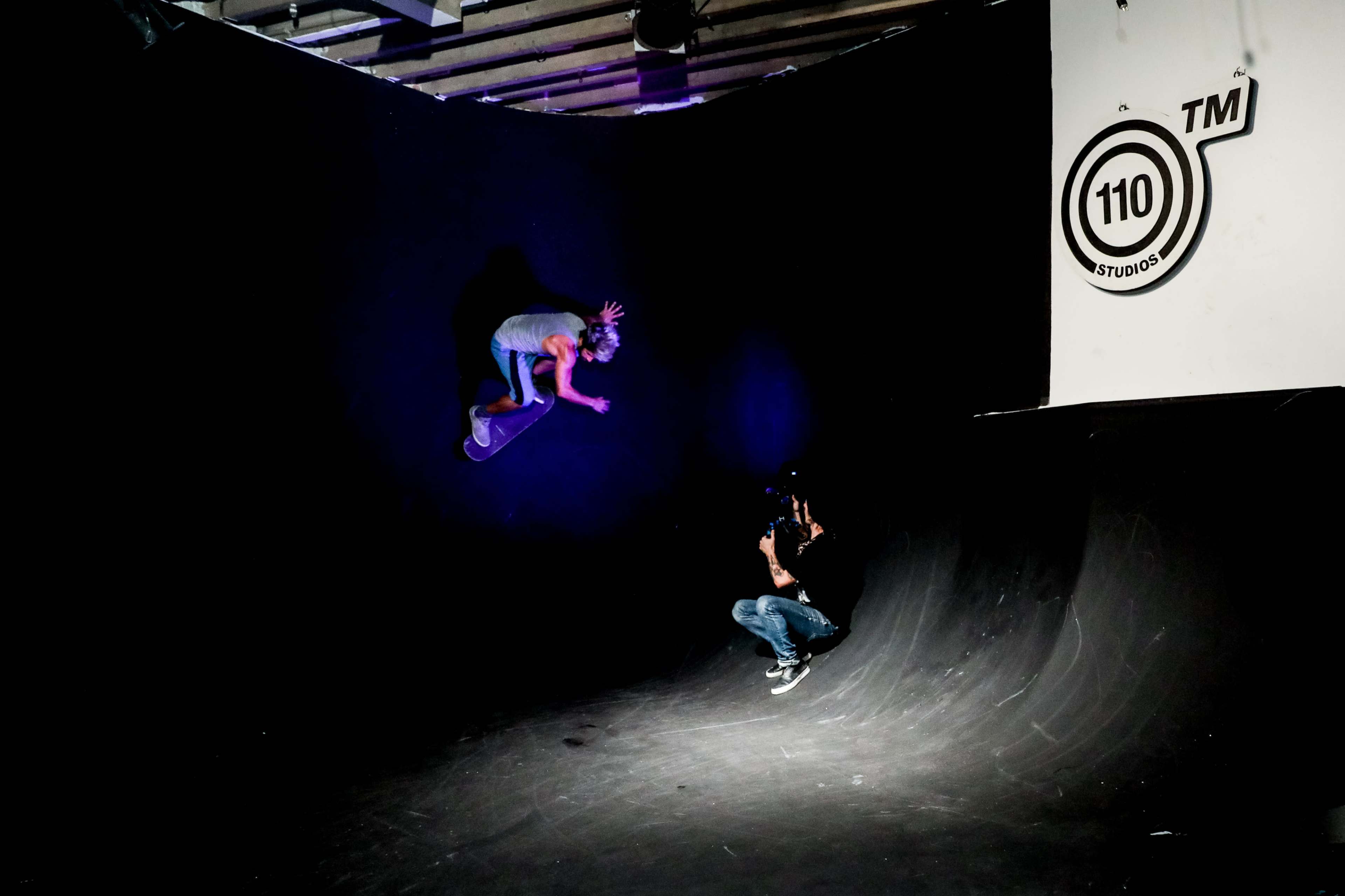 A skateboarder performs a trick against a dark background while a photographer captures the moment.