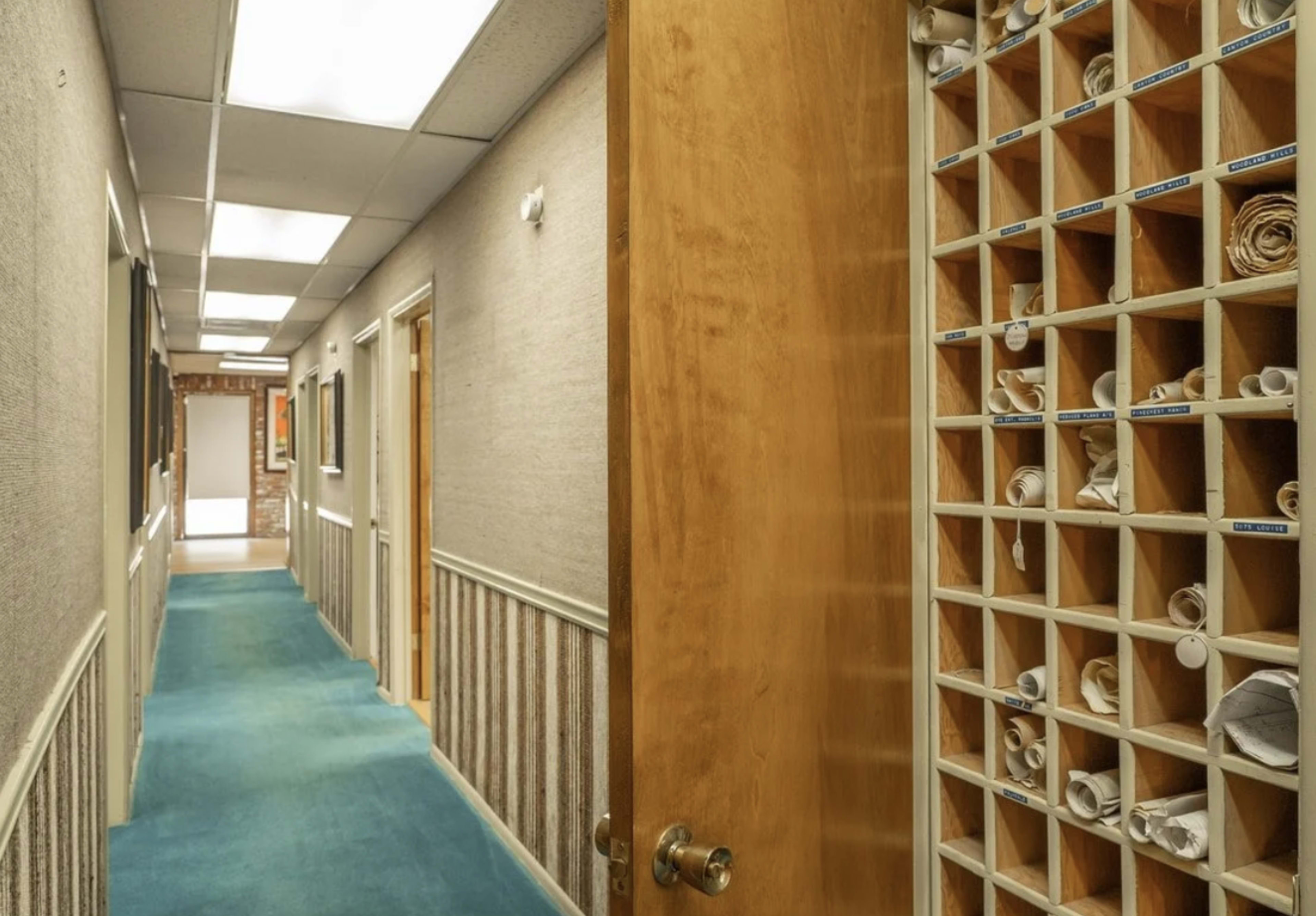 A wooden door opens to a narrow hallway lined with several doors and a shelving unit displaying rolled documents.