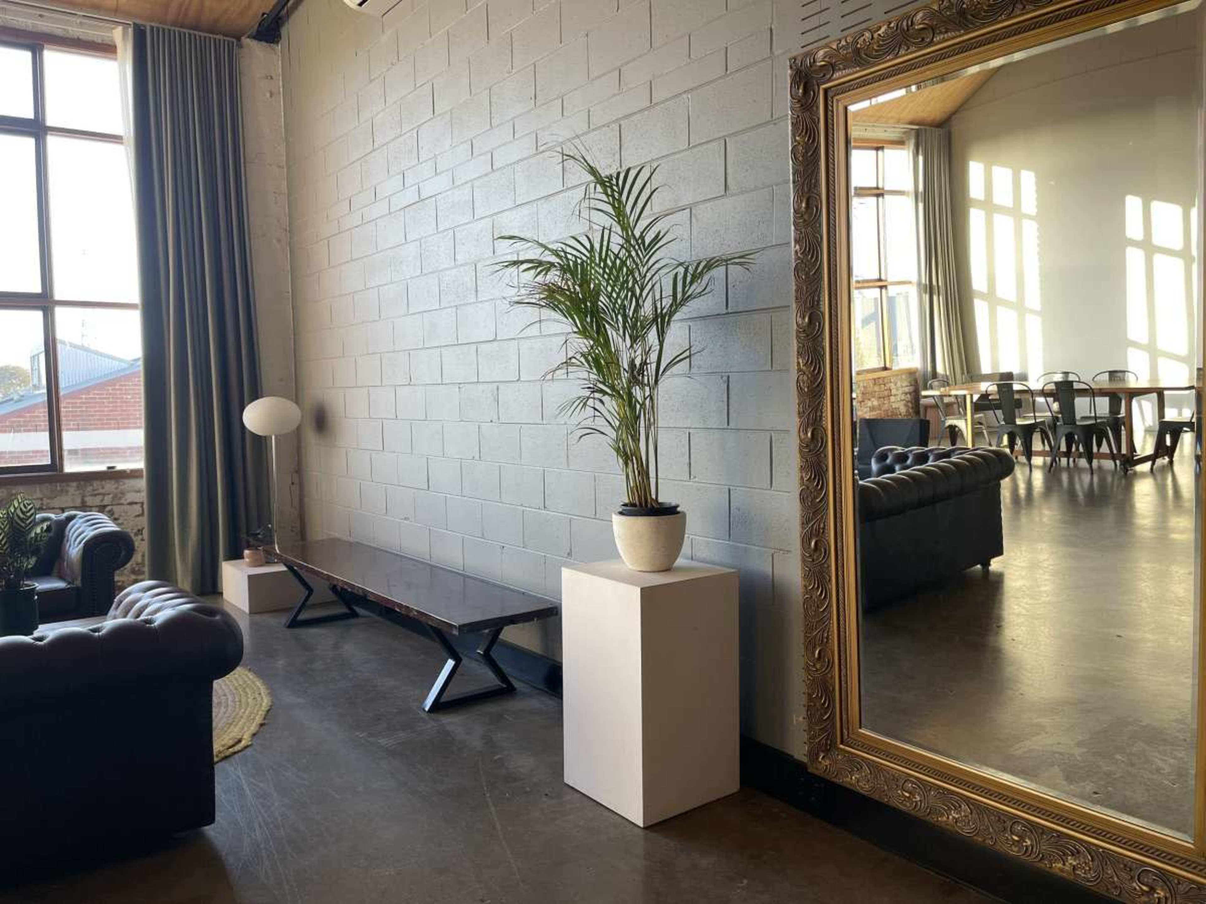 The image shows a modern interior space featuring a large mirror, a potted plant on a pedestal, a dark wood bench, and a seating area with a leather couch.