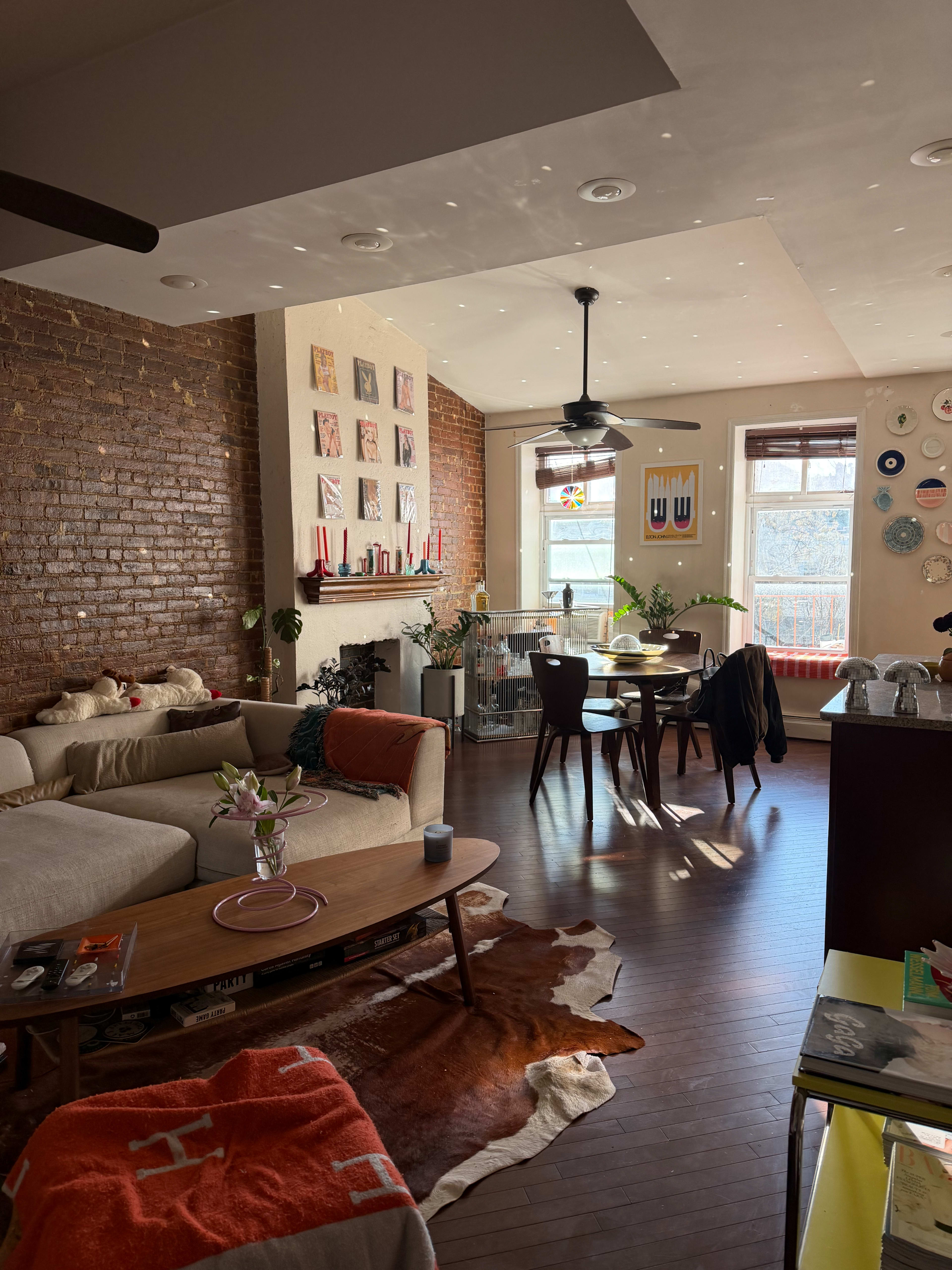 The image shows a bright and modern living space with exposed brick walls, a sectional sofa, a wooden coffee table, and a dining area with large windows.