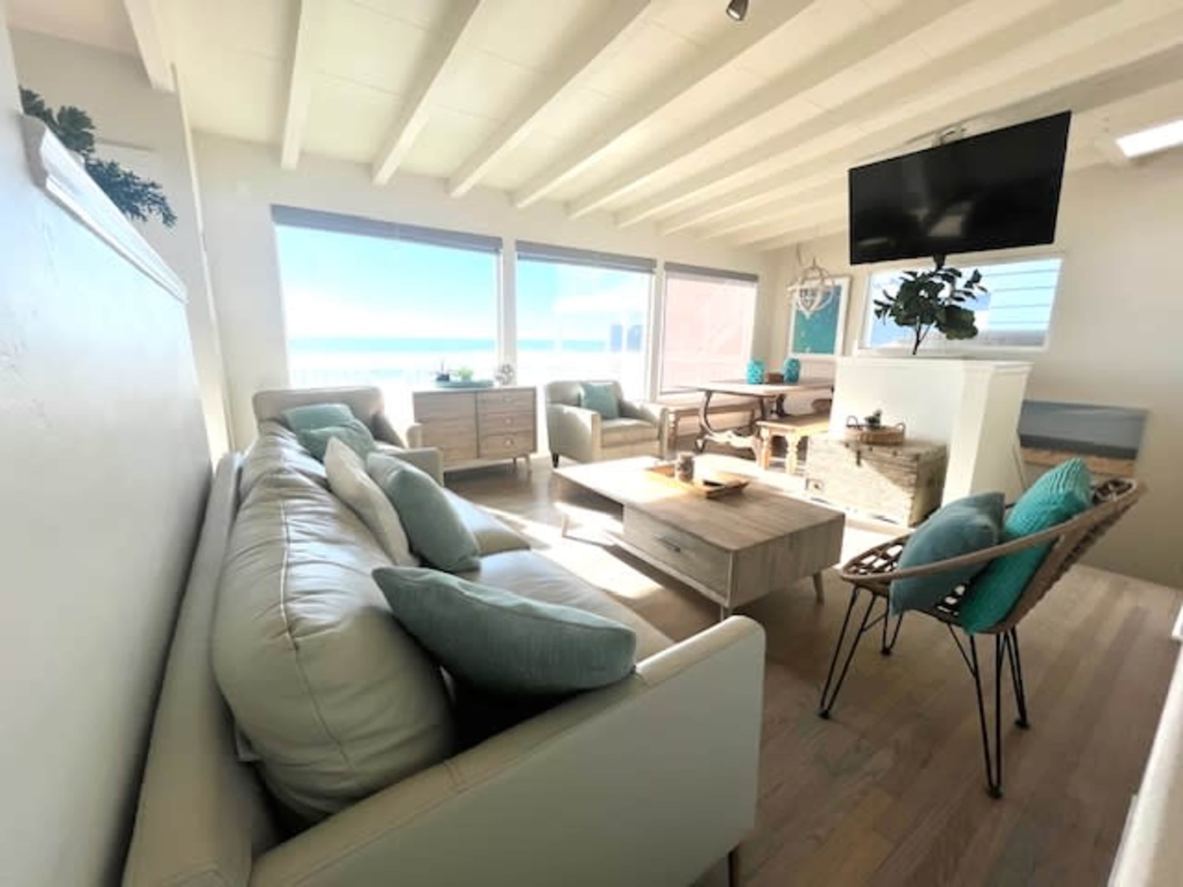 A bright living room features a light-colored sofa, a coffee table, and a large television, with windows offering a view of the ocean.