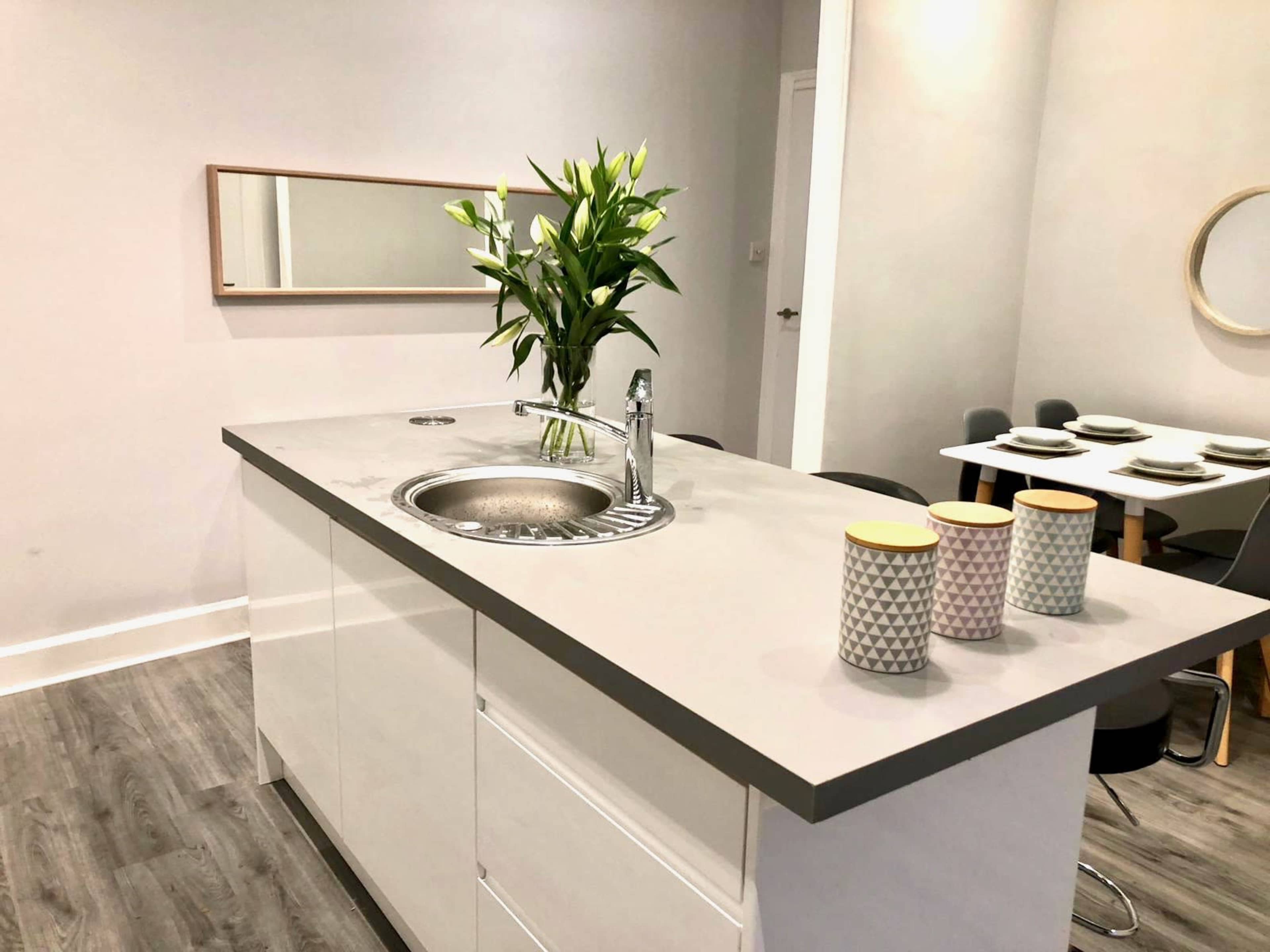 A modern kitchen features a central island with a sink, a vase of flowers, and decorative cups, with a dining area visible in the background.