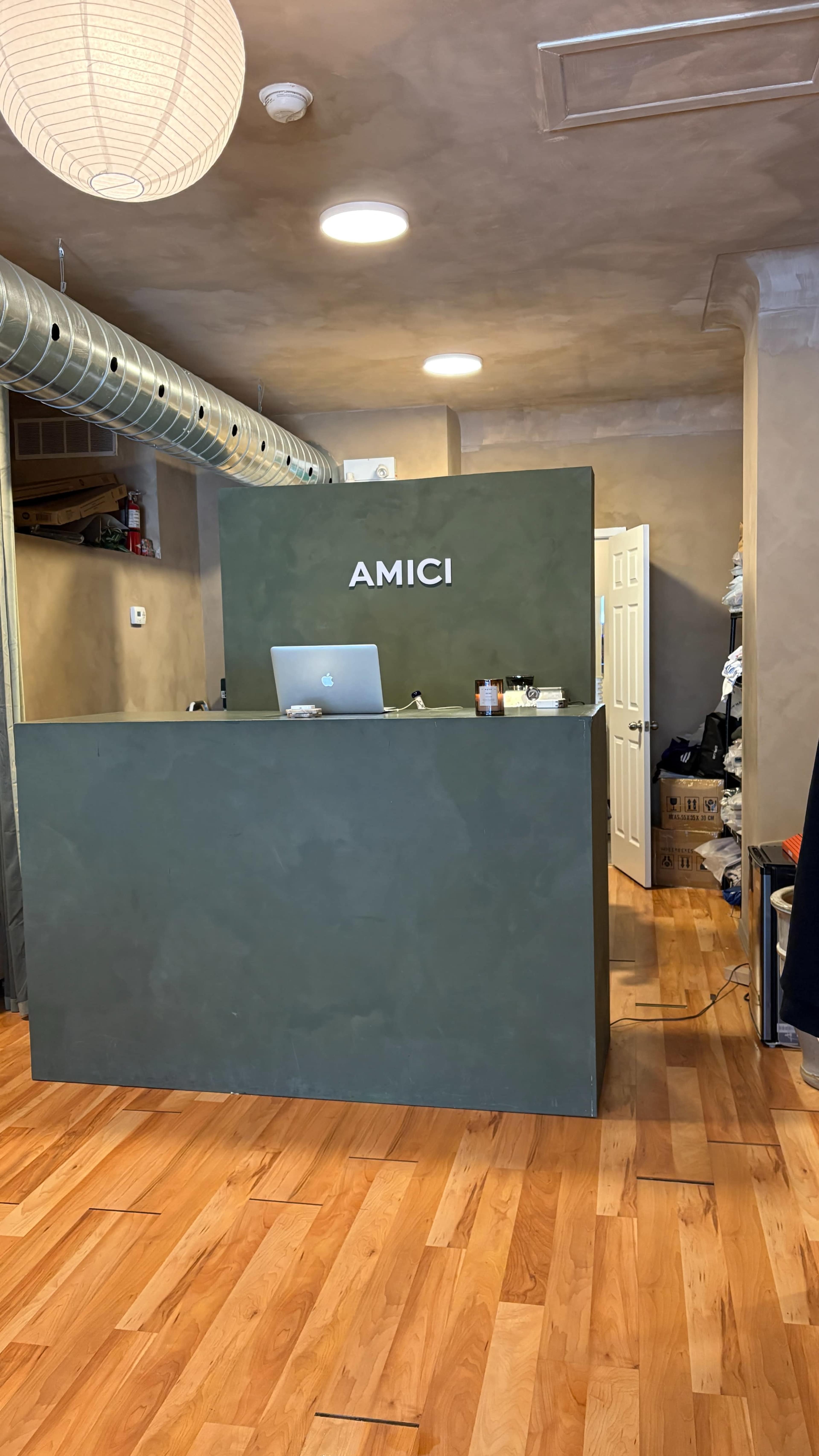 The image shows a reception area with a green desk labeled "AMICI," a computer on the desk, and a door leading to another room in the background.