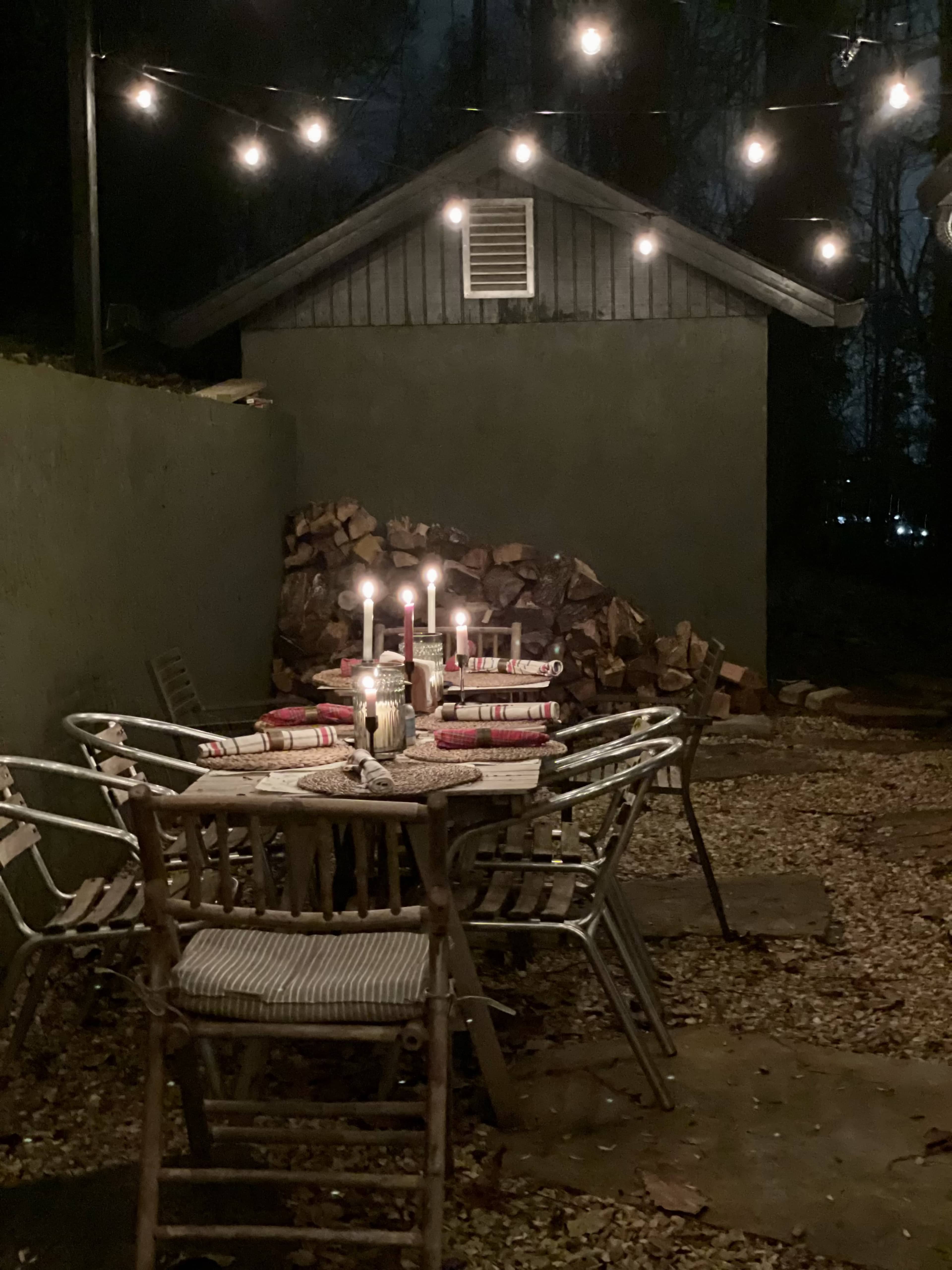 A dimly lit outdoor dining area features a table set with candles and surrounded by chairs, with a stack of firewood and string lights in the background.