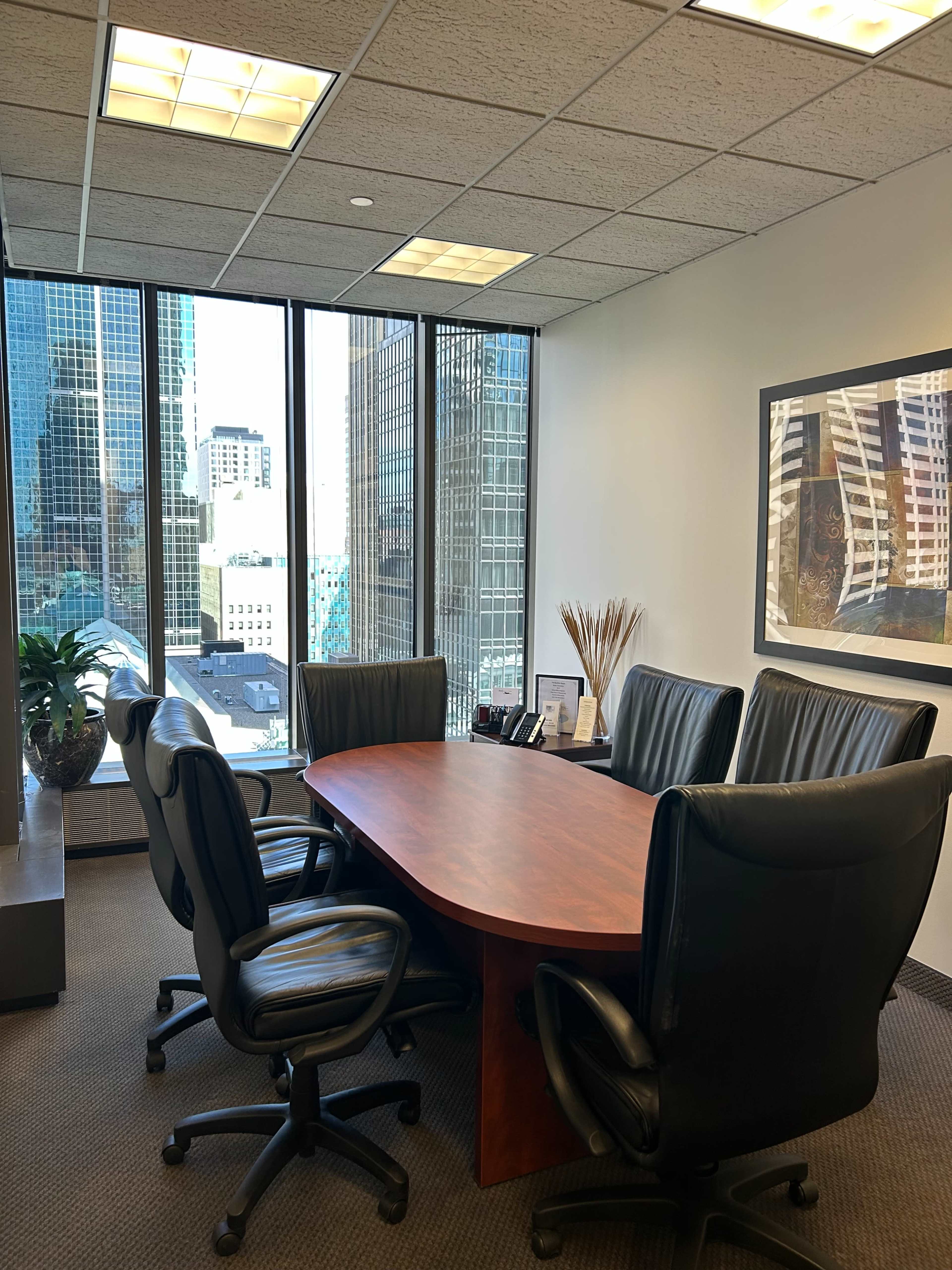 A conference room features a large oval table surrounded by five black leather chairs, with large windows offering a view of city skyscrapers.