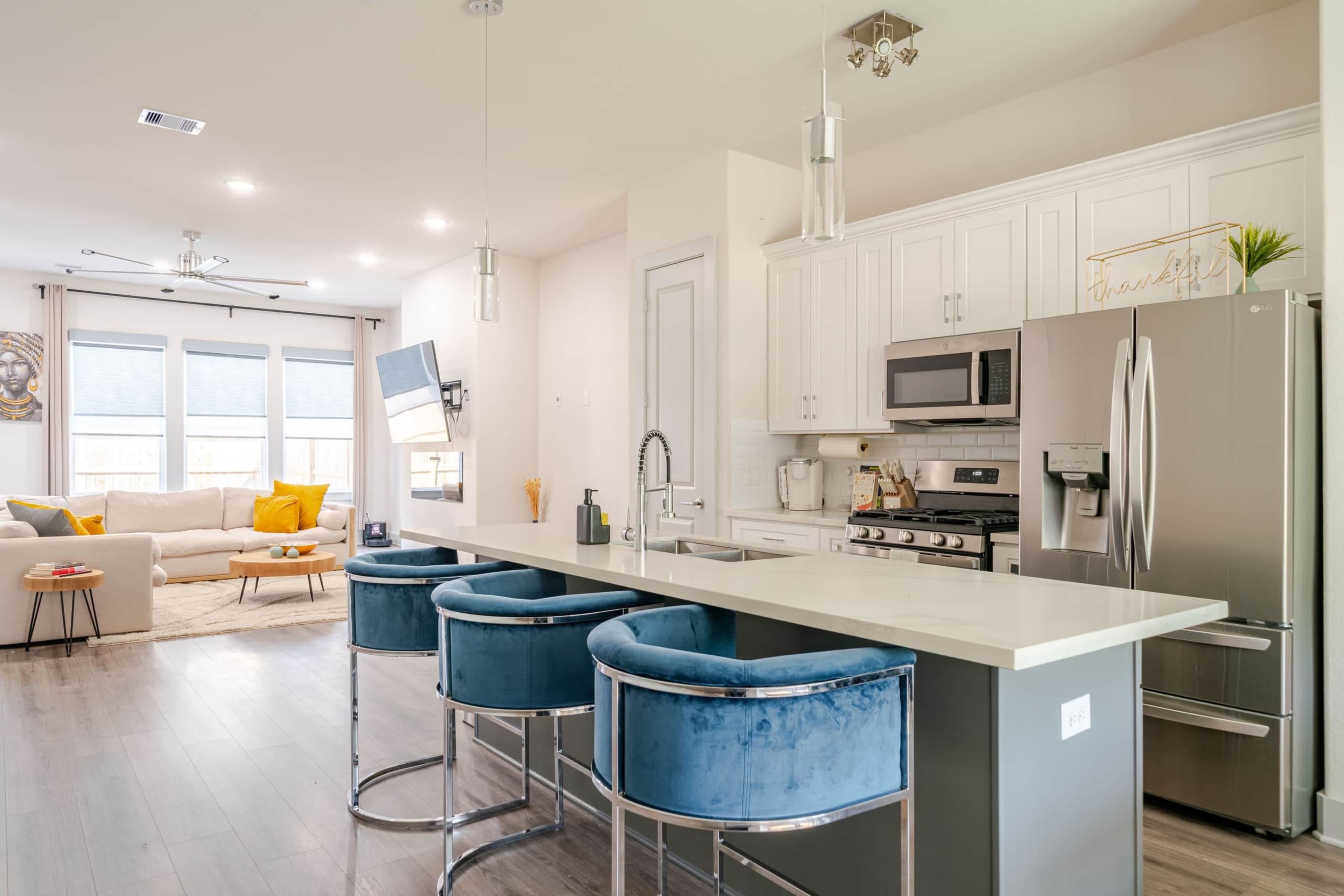 The image shows a modern kitchen with white cabinetry, stainless steel appliances, and blue barstools at a counter, overlooking a bright living area.