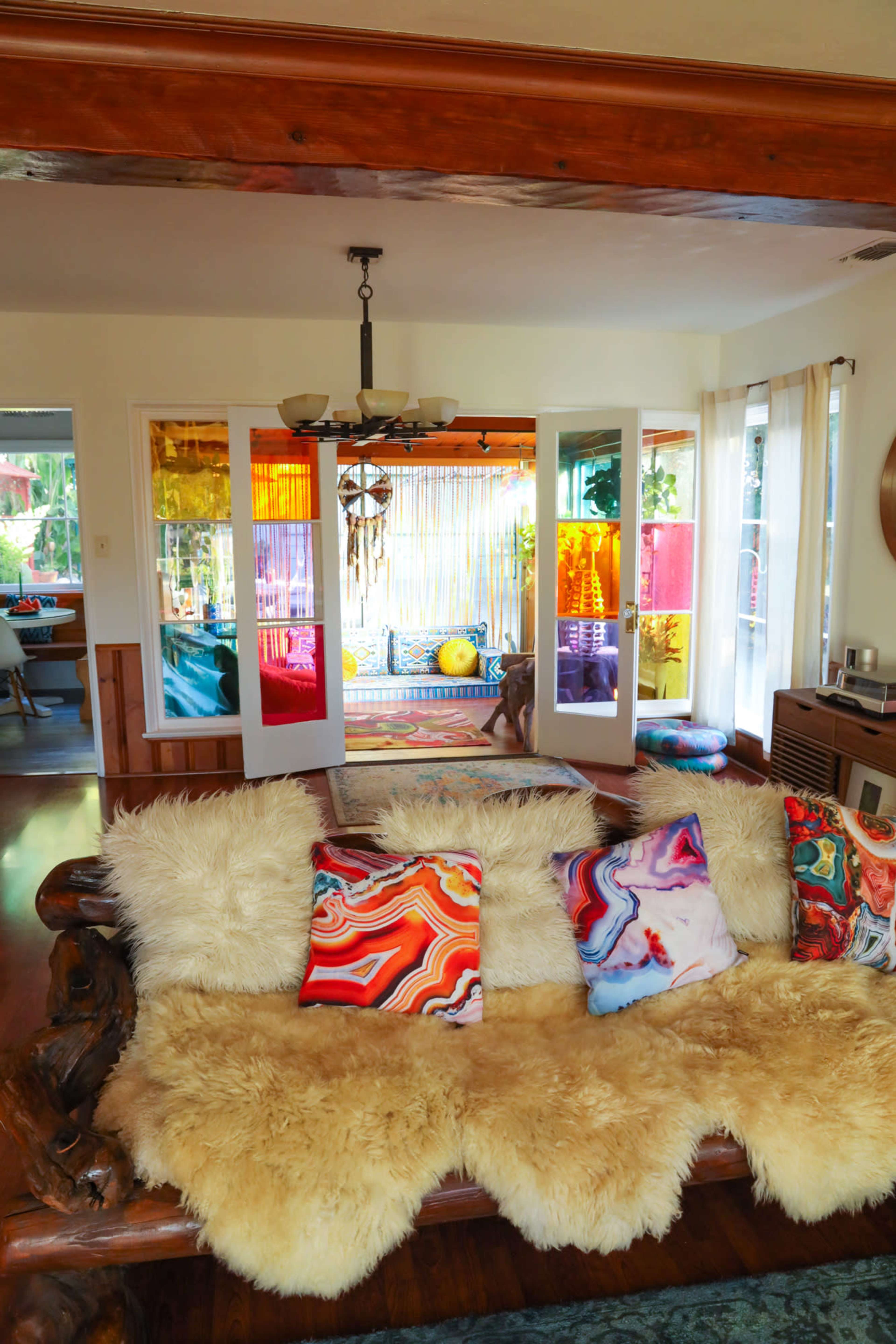 A wooden sofa with fur throws and colorful patterned pillows sits facing open doors leading to a bright, vibrant room decorated with hanging curtains and cushions.