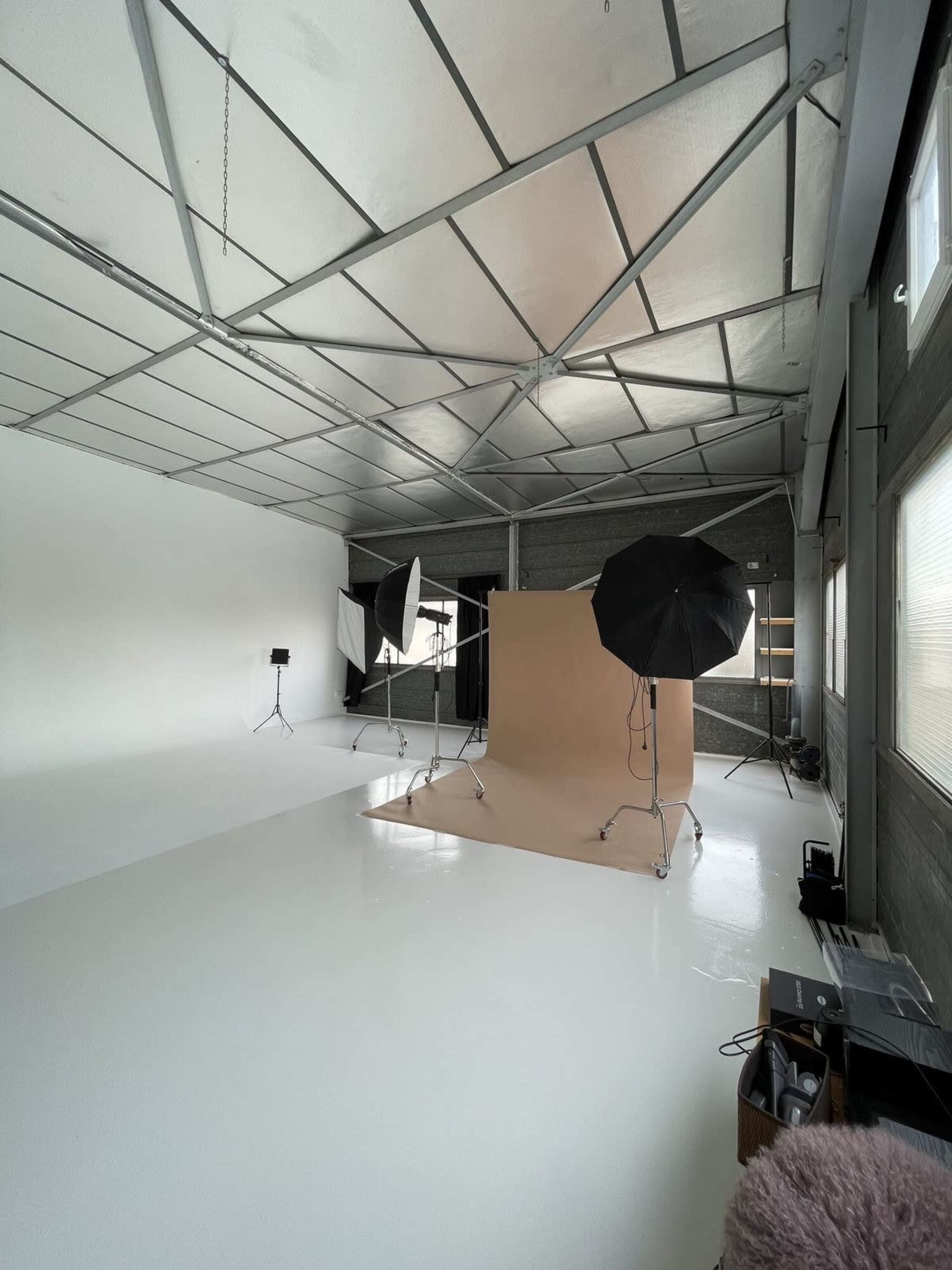 The image shows a spacious photography studio with a beige backdrop, three lighting setups, and white polished floors, all under a metal ceiling.