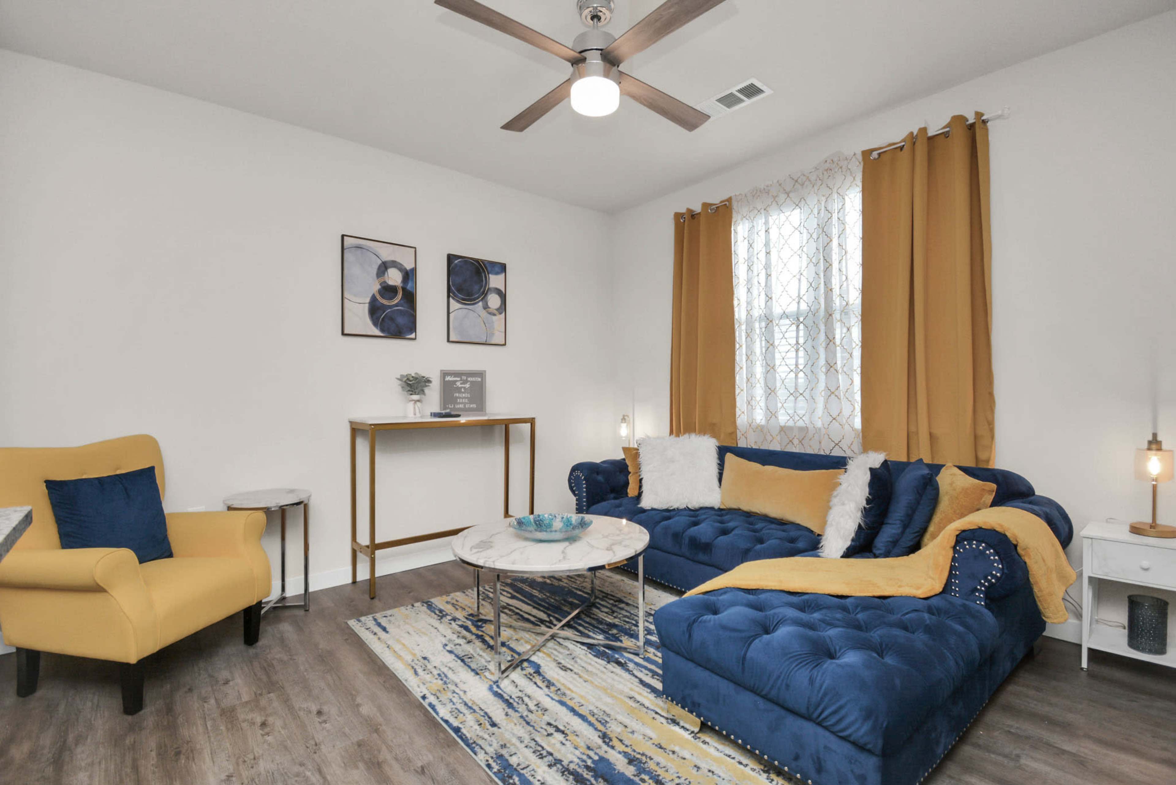 The living room features a blue sectional sofa, a round marble coffee table, a yellow armchair, and large yellow curtains.