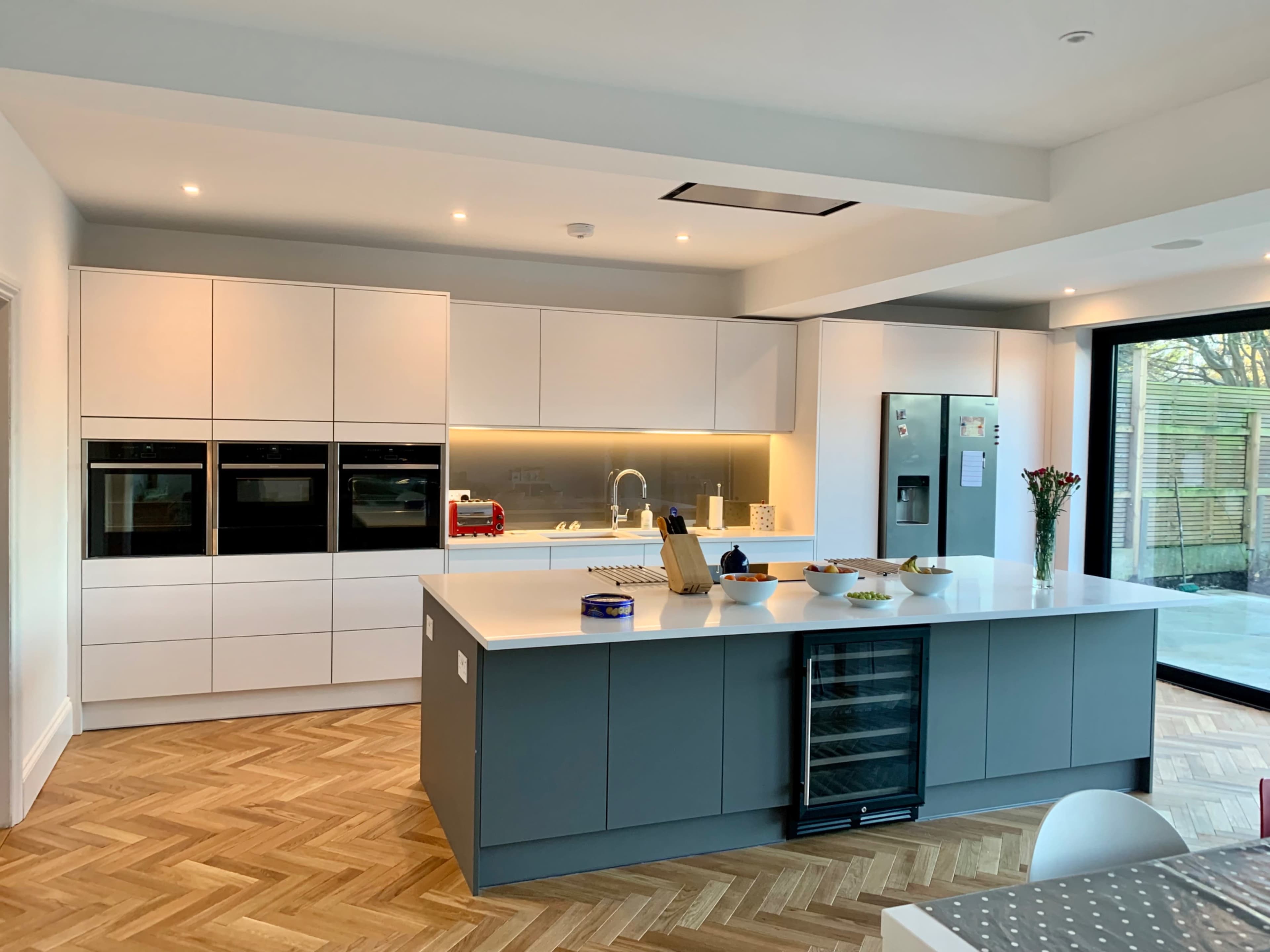 A modern kitchen with a central island, equipped with built-in ovens, a refrigerator, and a mix of white and gray cabinetry, all set against a wooden herringbone floor.