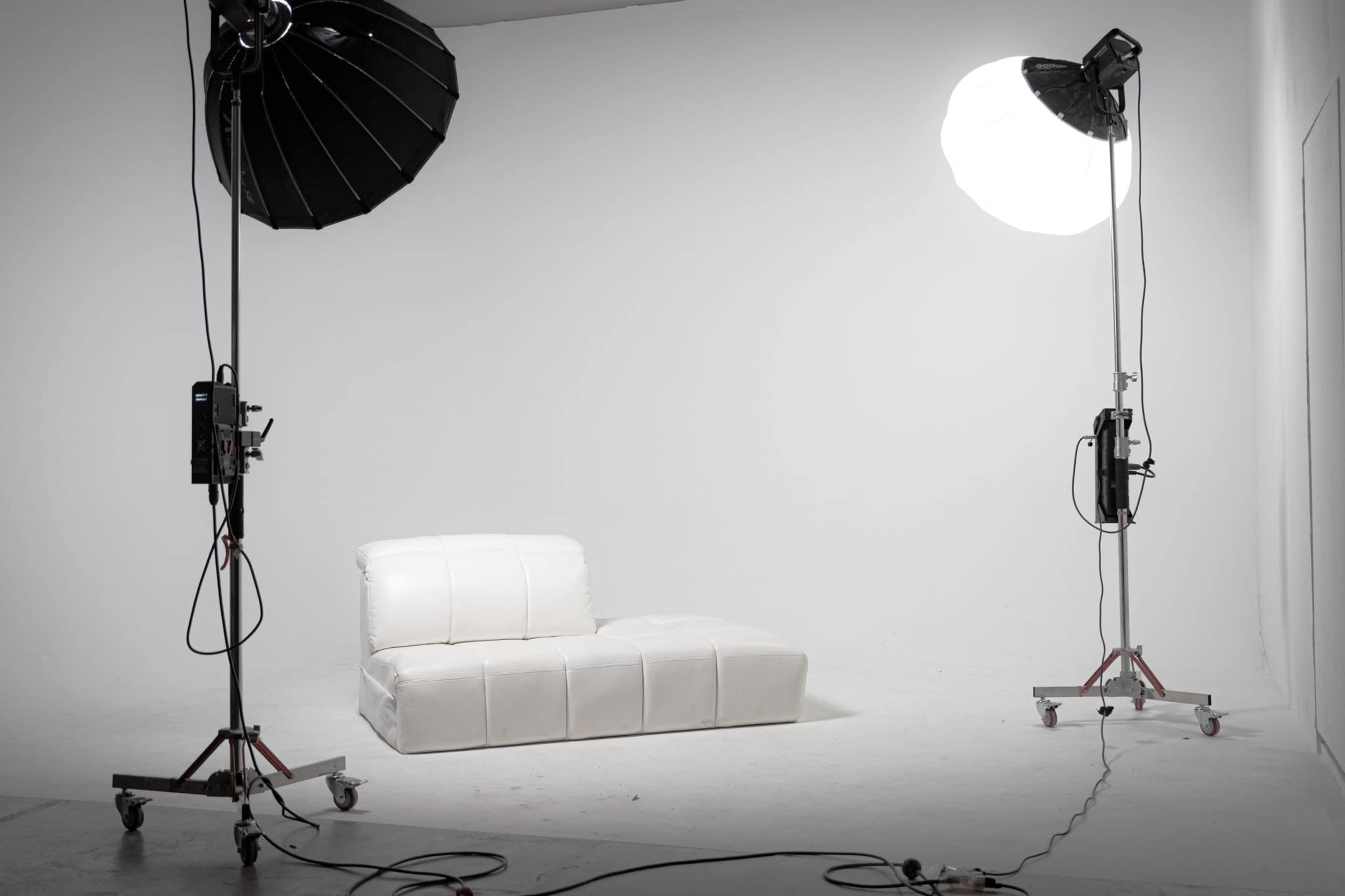 A white sofa is positioned between two studio lights in a minimalist photography studio.