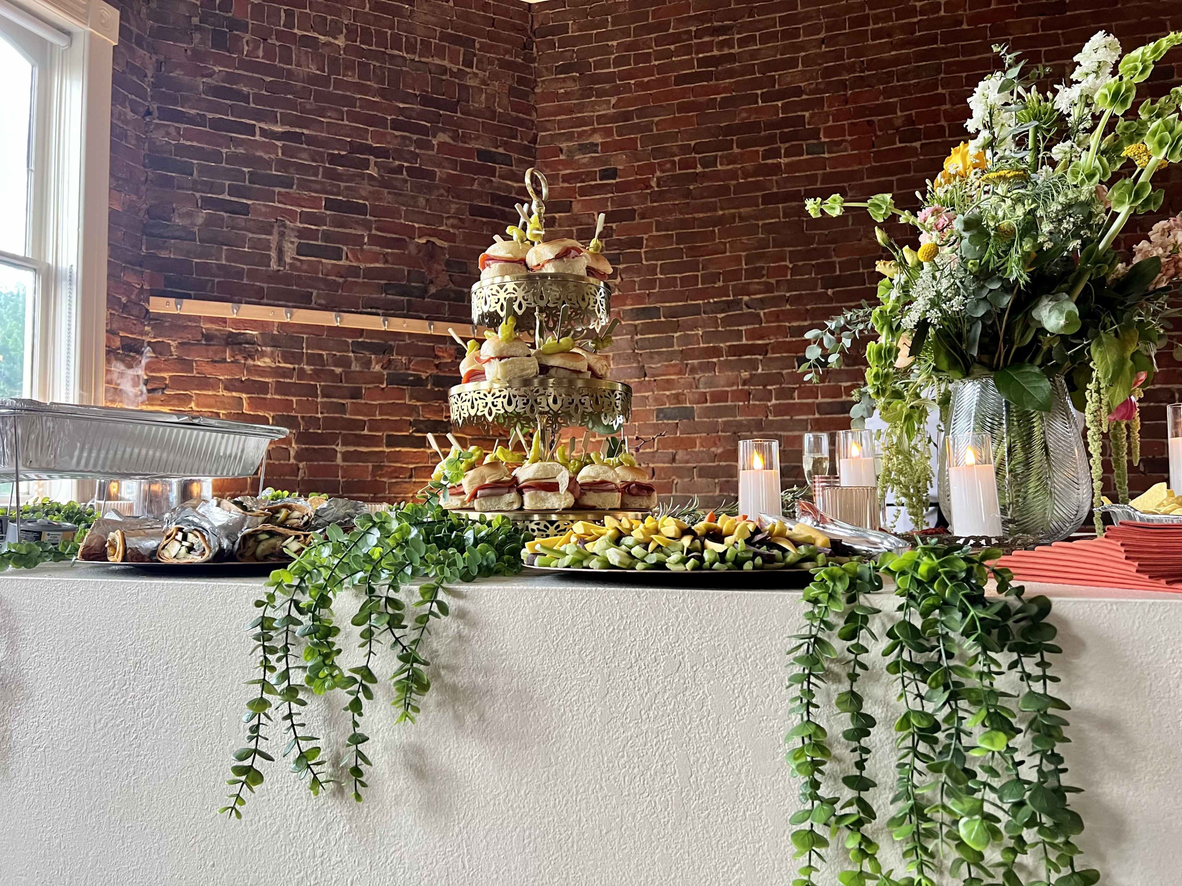 A decorative food display featuring a multi-tiered platter of sandwiches, surrounded by various appetizers and candles, on a table adorned with greenery against a brick wall.