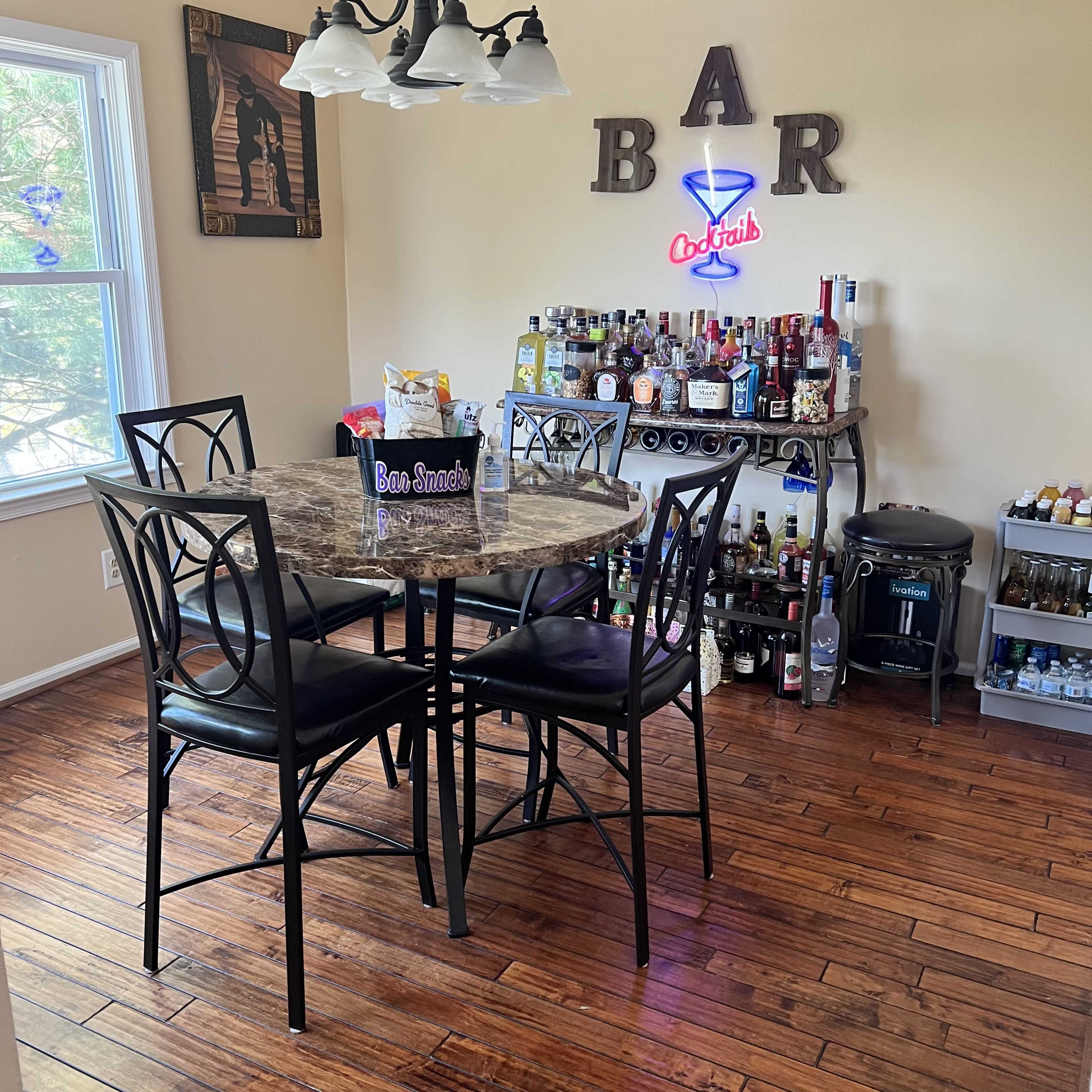 A dining area features a round marble table with four black chairs and a bar cart stocked with various bottles and a neon sign above.