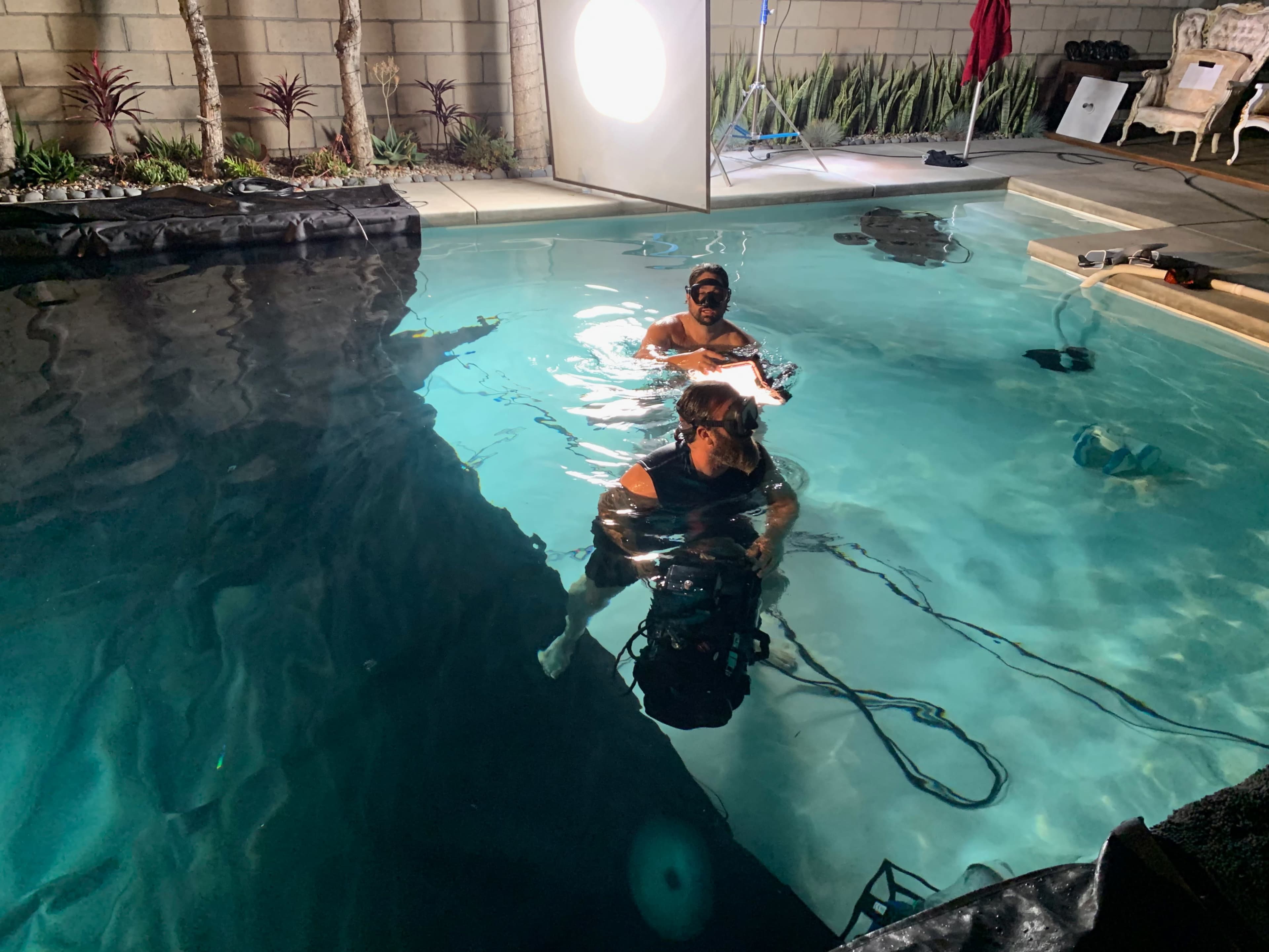 Two individuals are submerged in a pool while filming, with equipment positioned around the water.