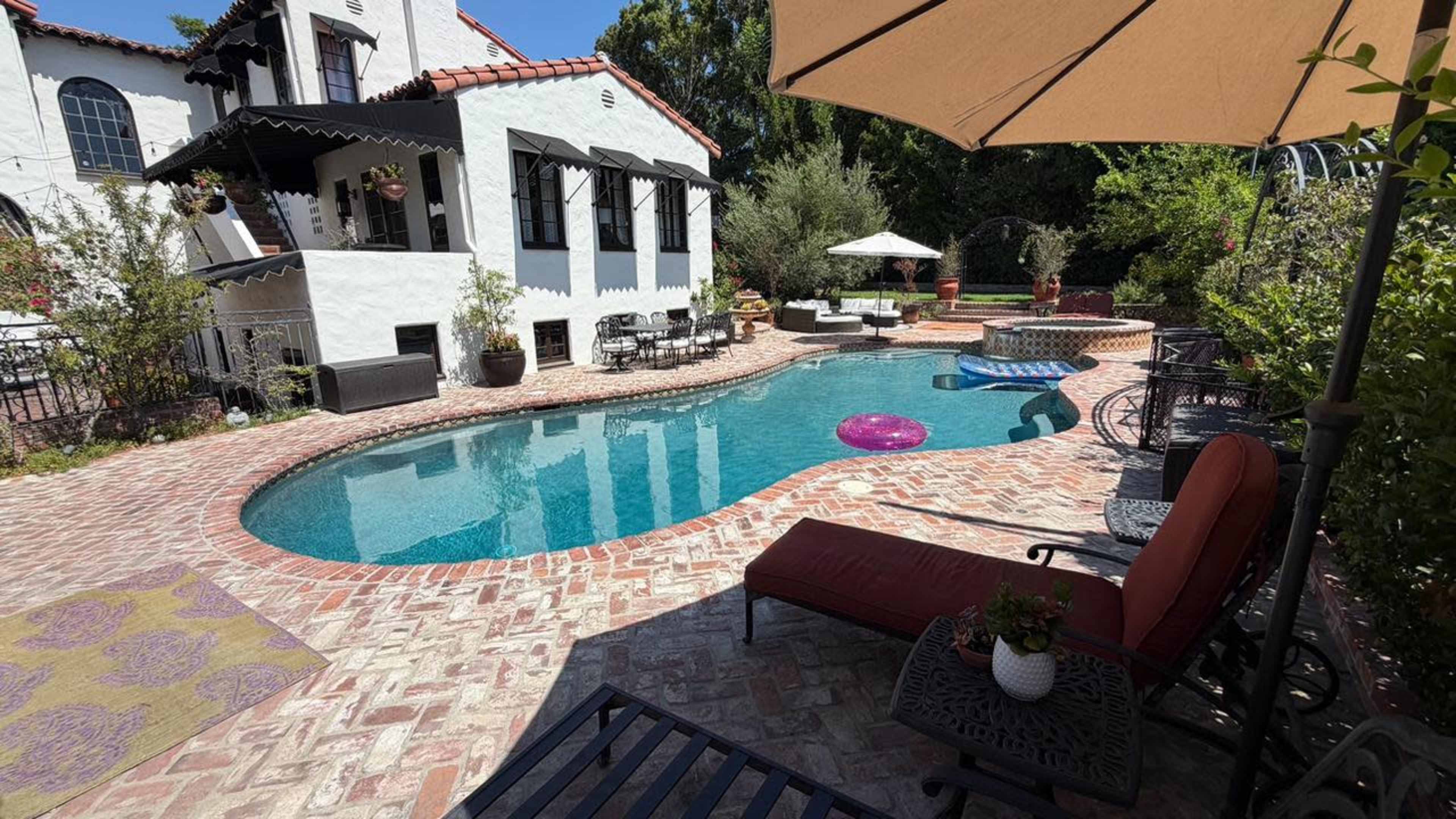 The image shows a residential backyard with a swimming pool surrounded by brick patios, lounge chairs, and pool floats, complemented by lush greenery and outdoor seating areas.