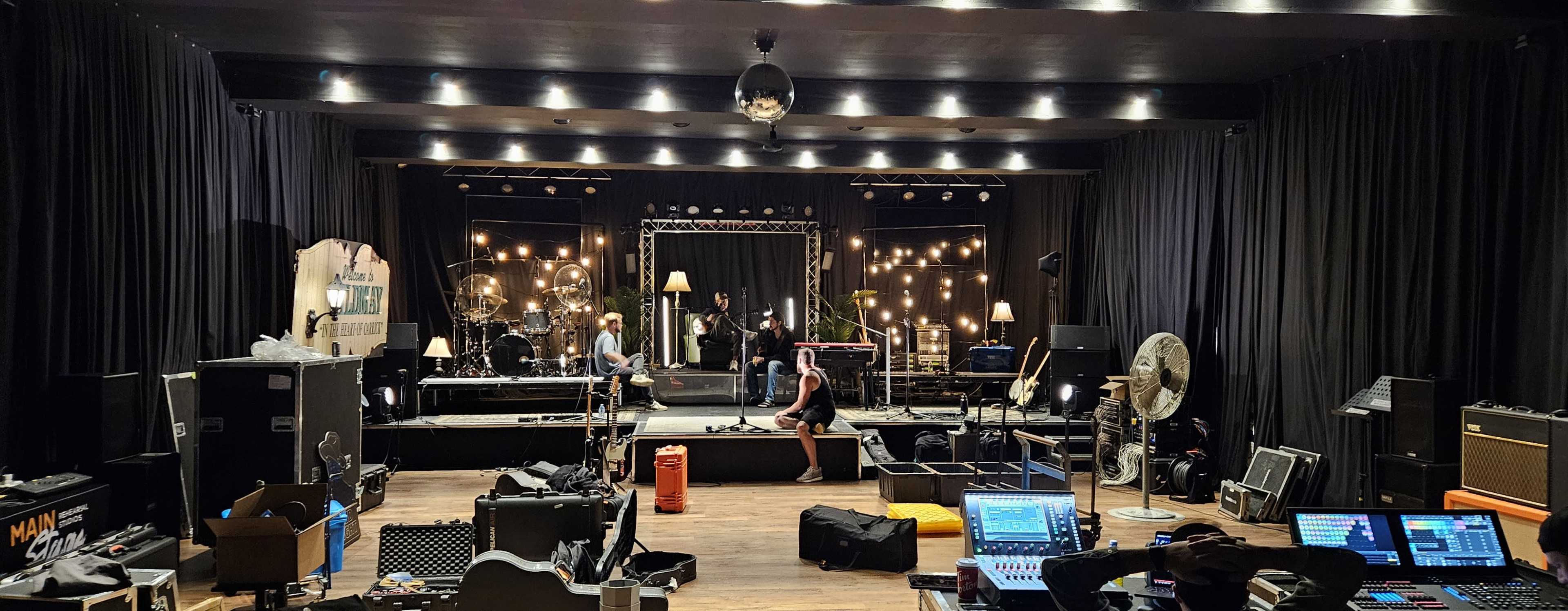 The image shows a dimly lit stage with a band setup, sound equipment, and scattered boxes in a music venue.