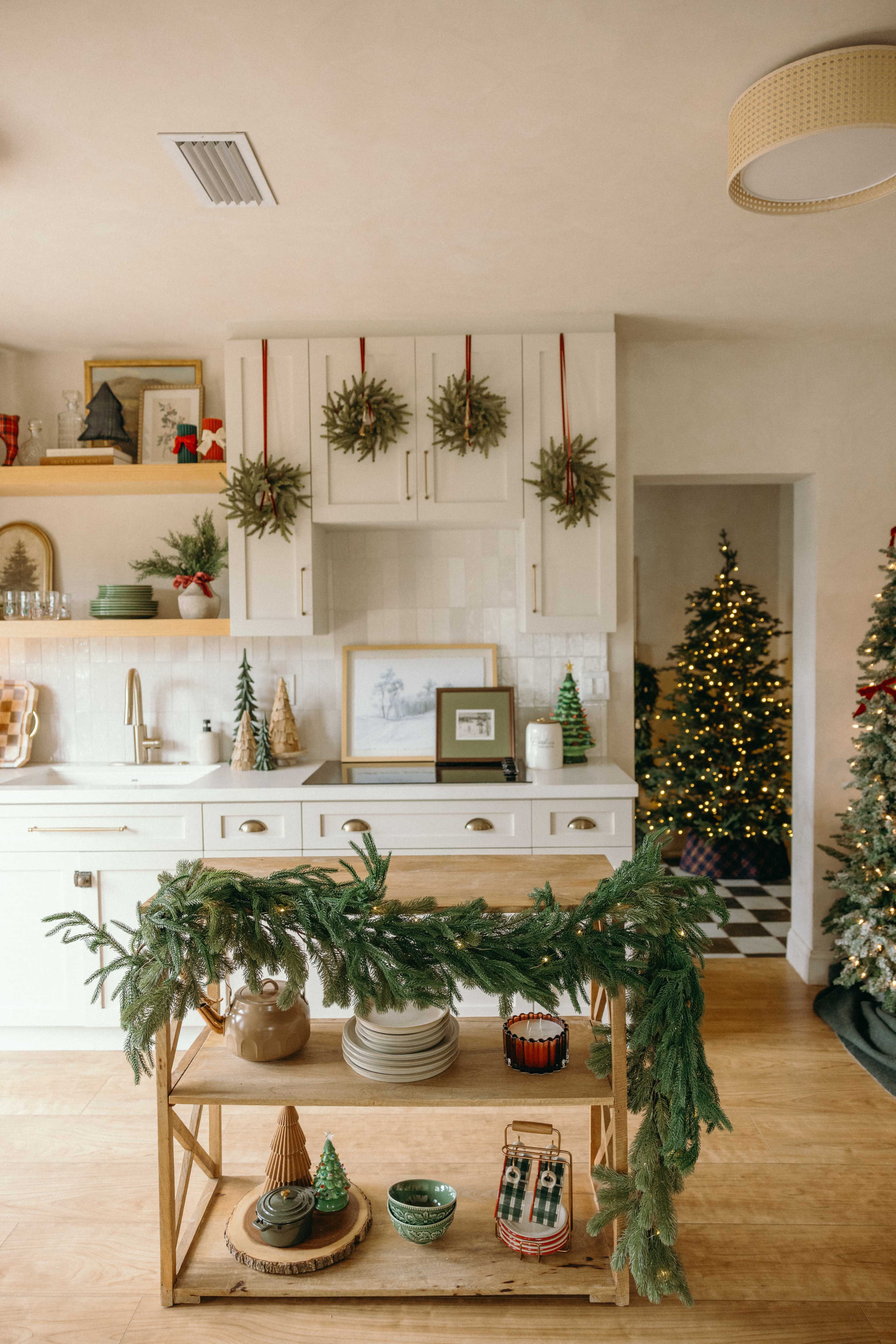The kitchen features a wooden cart adorned with greenery, surrounded by festive decorations, while a Christmas tree illuminated with lights stands in the background.