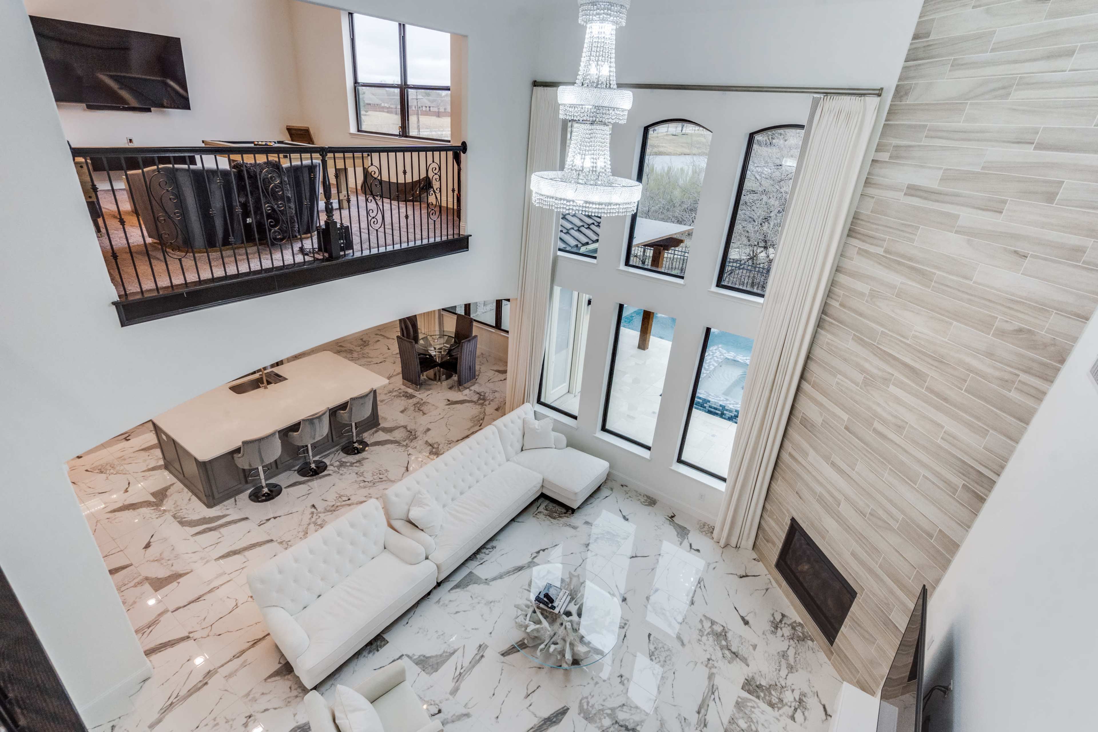 The image shows a large, modern living area with a white sectional sofa, high ceilings, large windows, and a crystal chandelier.