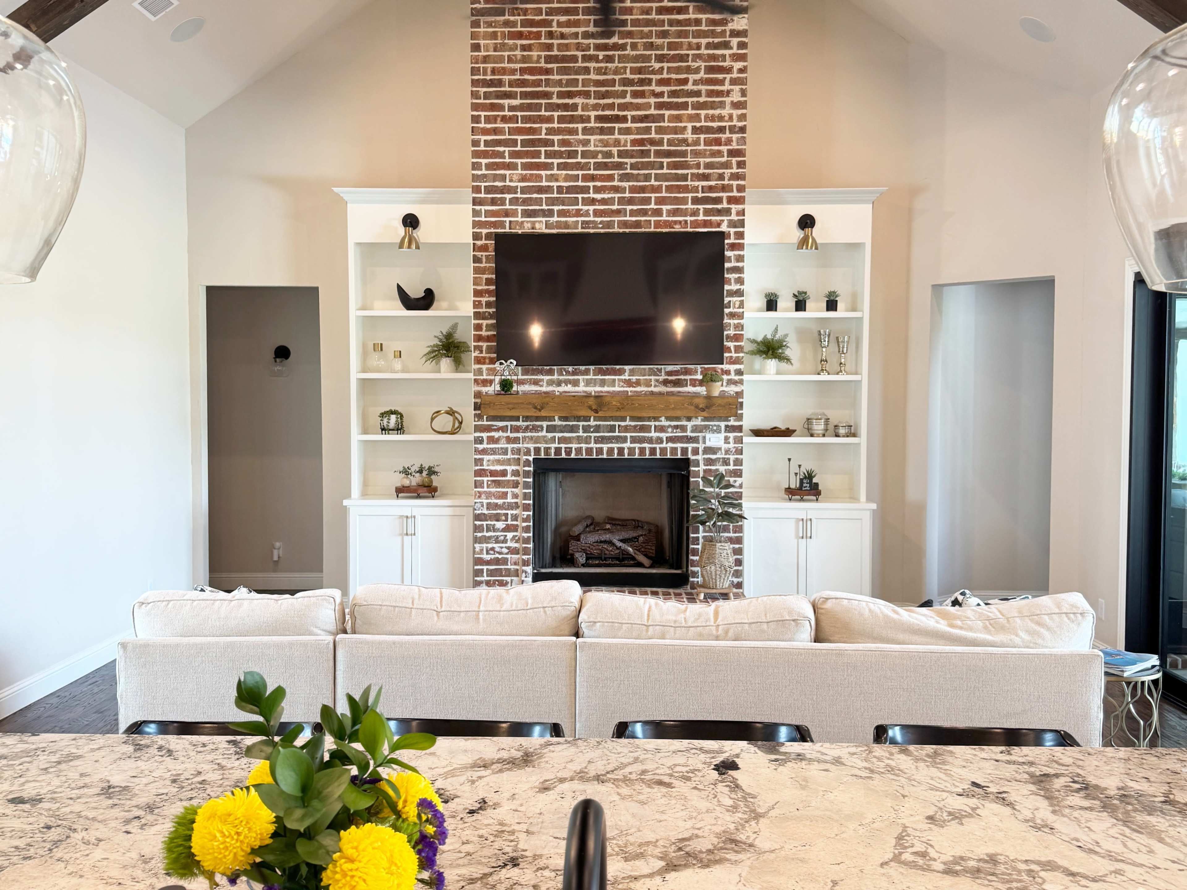 The image shows a living room with a brick fireplace, built-in shelves on either side, and a large television above the fireplace, all viewed from a kitchen with a countertop in the foreground.