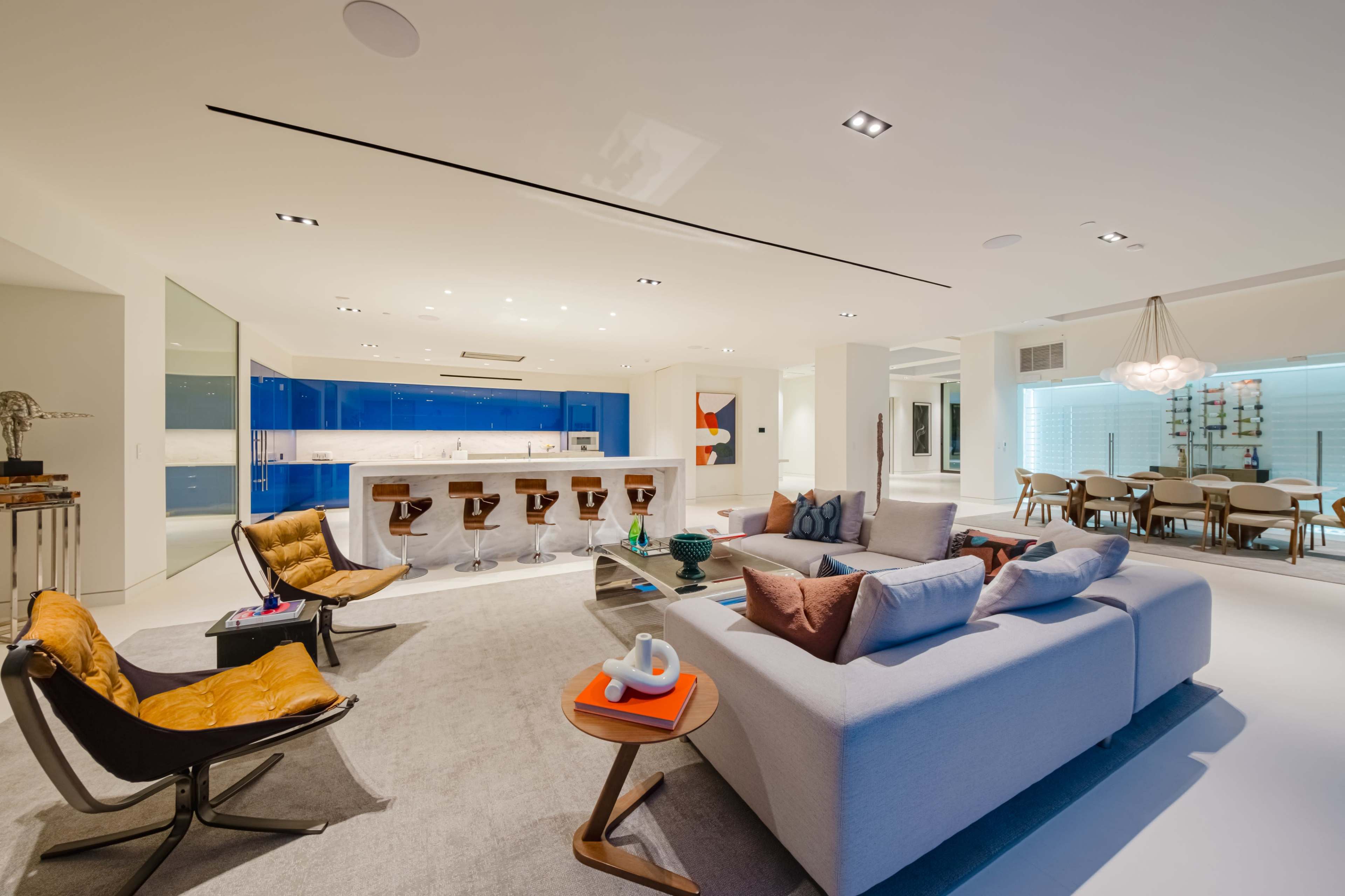 A modern open-concept living area featuring a gray sofa, contemporary chairs, a sleek kitchen with blue cabinets, and a dining area with a large table.