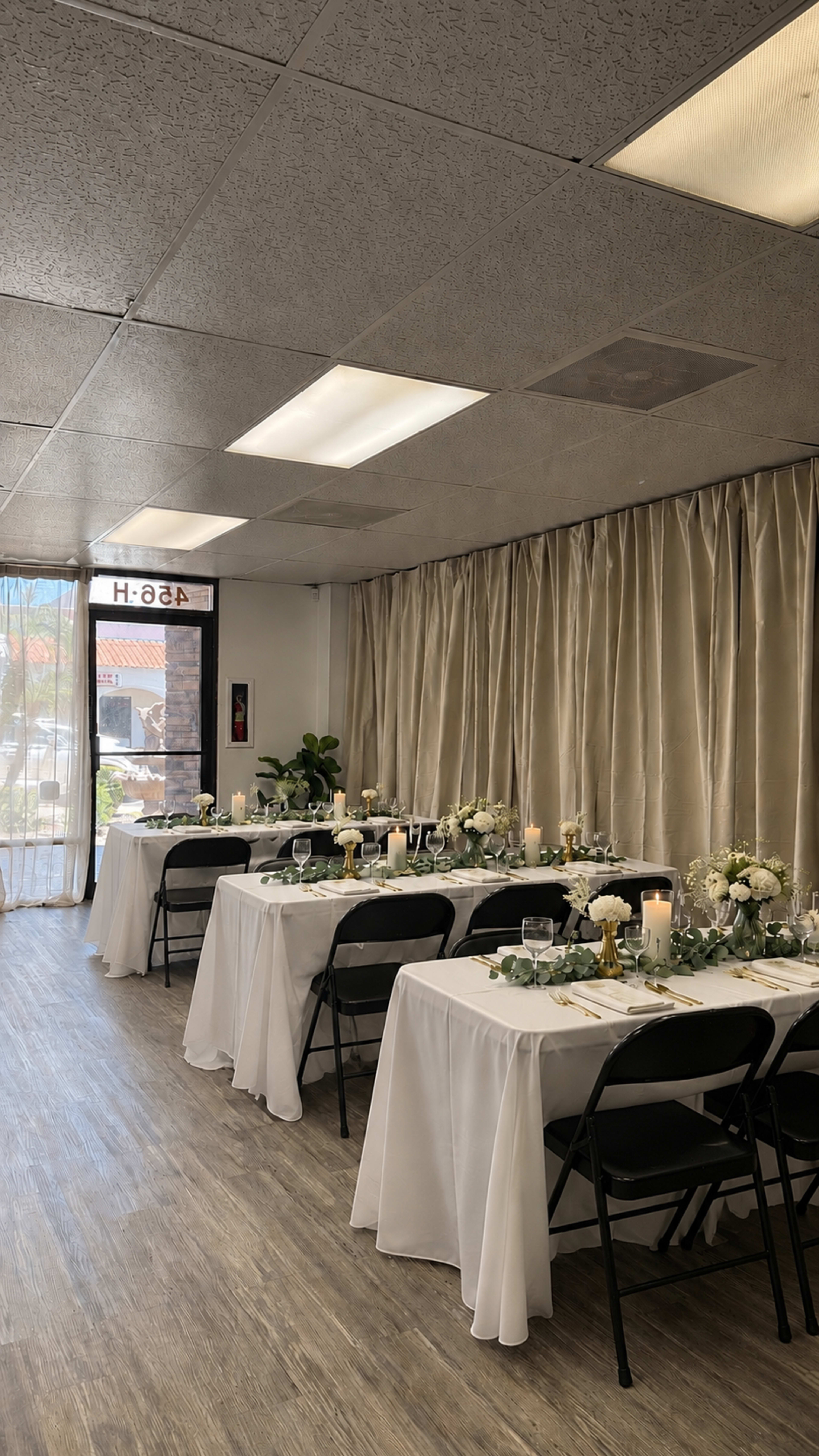 The image shows a decorated event space with neatly arranged tables covered in white tablecloths, set for a gathering, and flanked by beige curtains and large windows.