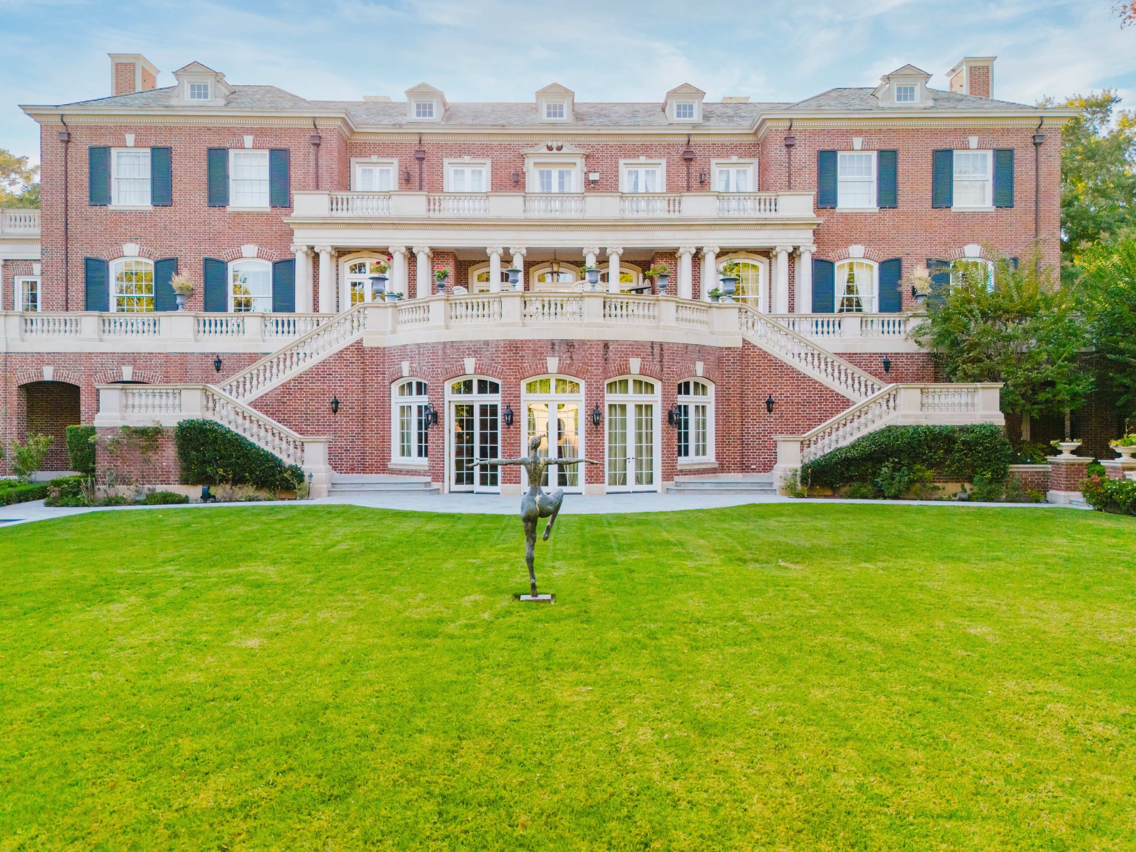 A large brick mansion features a grand facade with multiple balconies, expansive staircases, and a neatly landscaped lawn with a sculpture in the foreground.