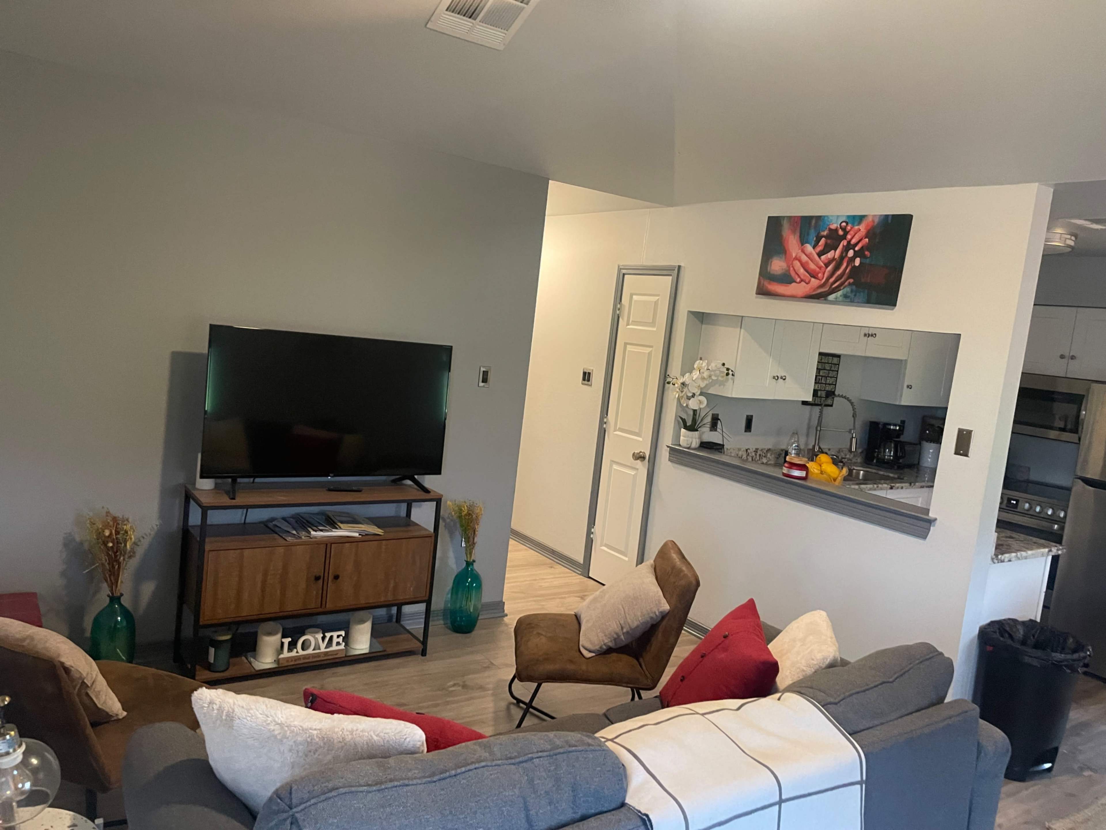 The image shows a modern living room with a gray sofa, a wooden TV stand, and a kitchen area visible in the background.