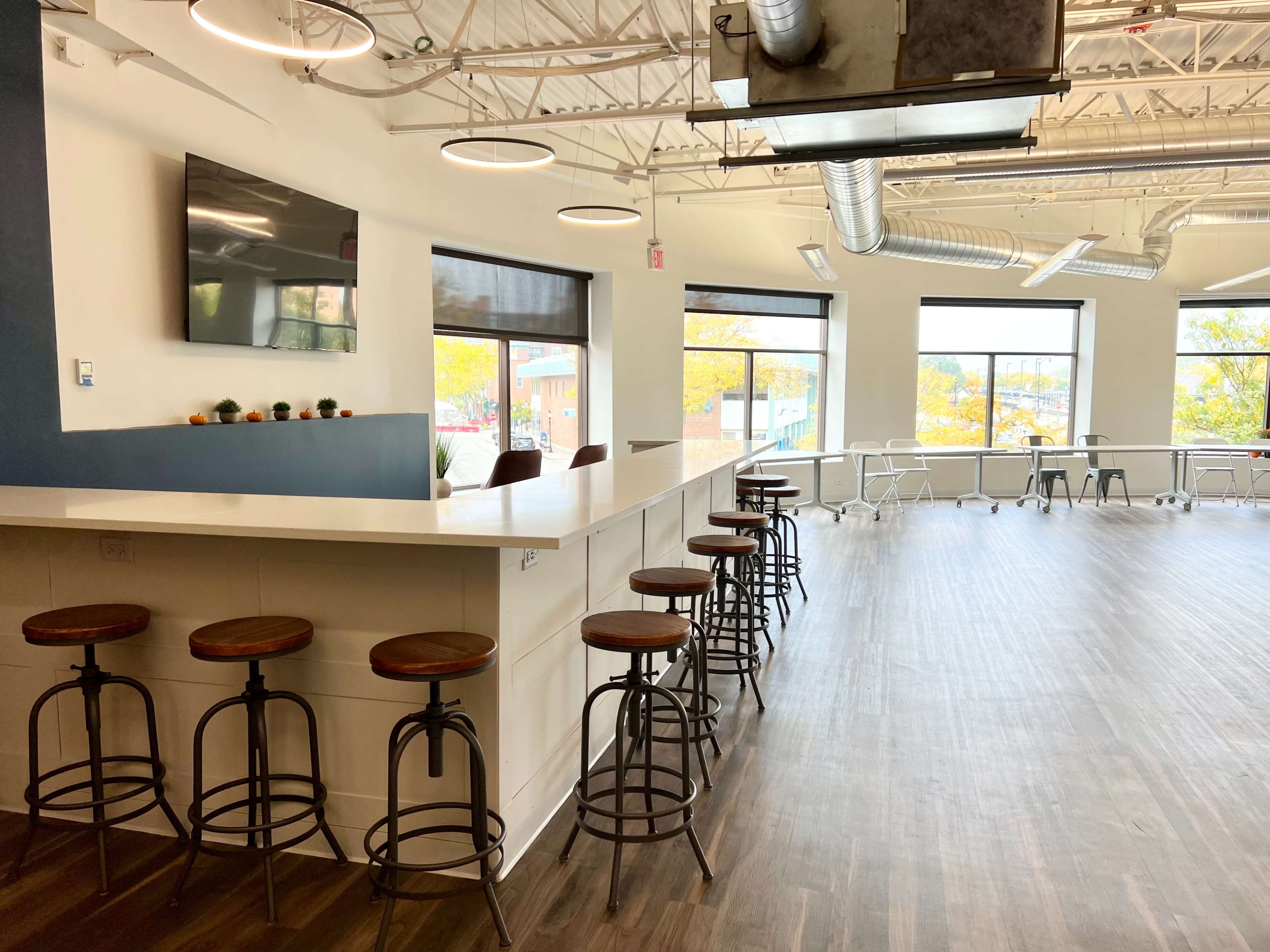 A modern office interior with a bar-height counter and several stools on one side, complemented by large windows letting in natural light.