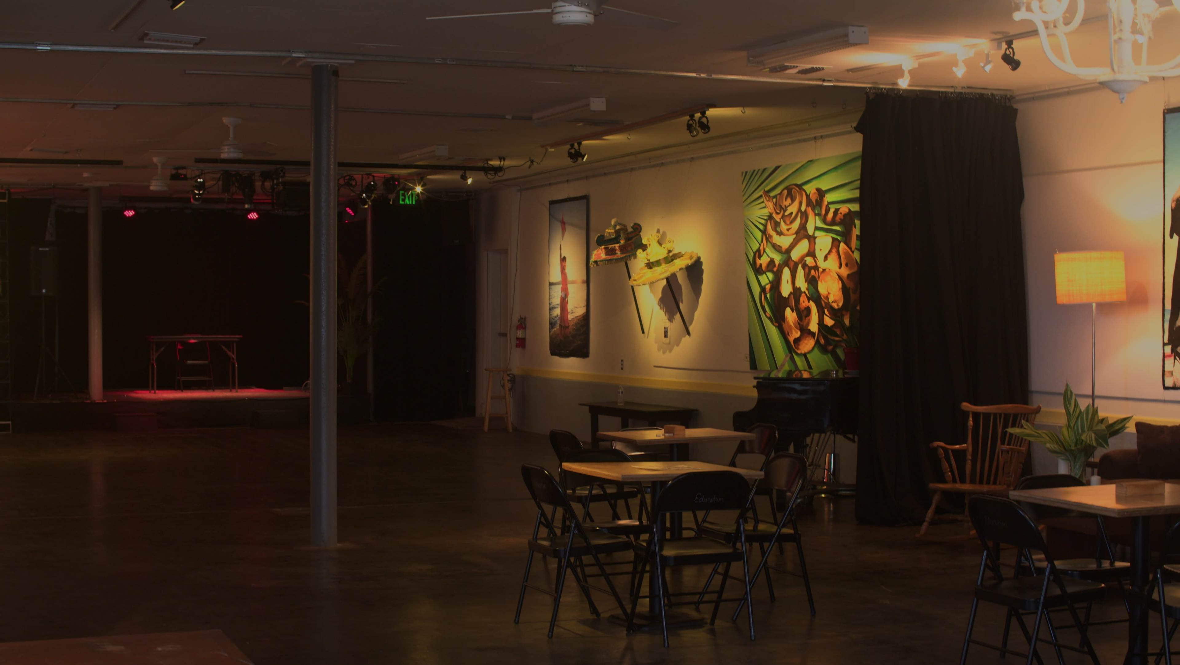 The image shows an empty venue with tables and chairs arranged in front of a stage adorned with colorful wall art.
