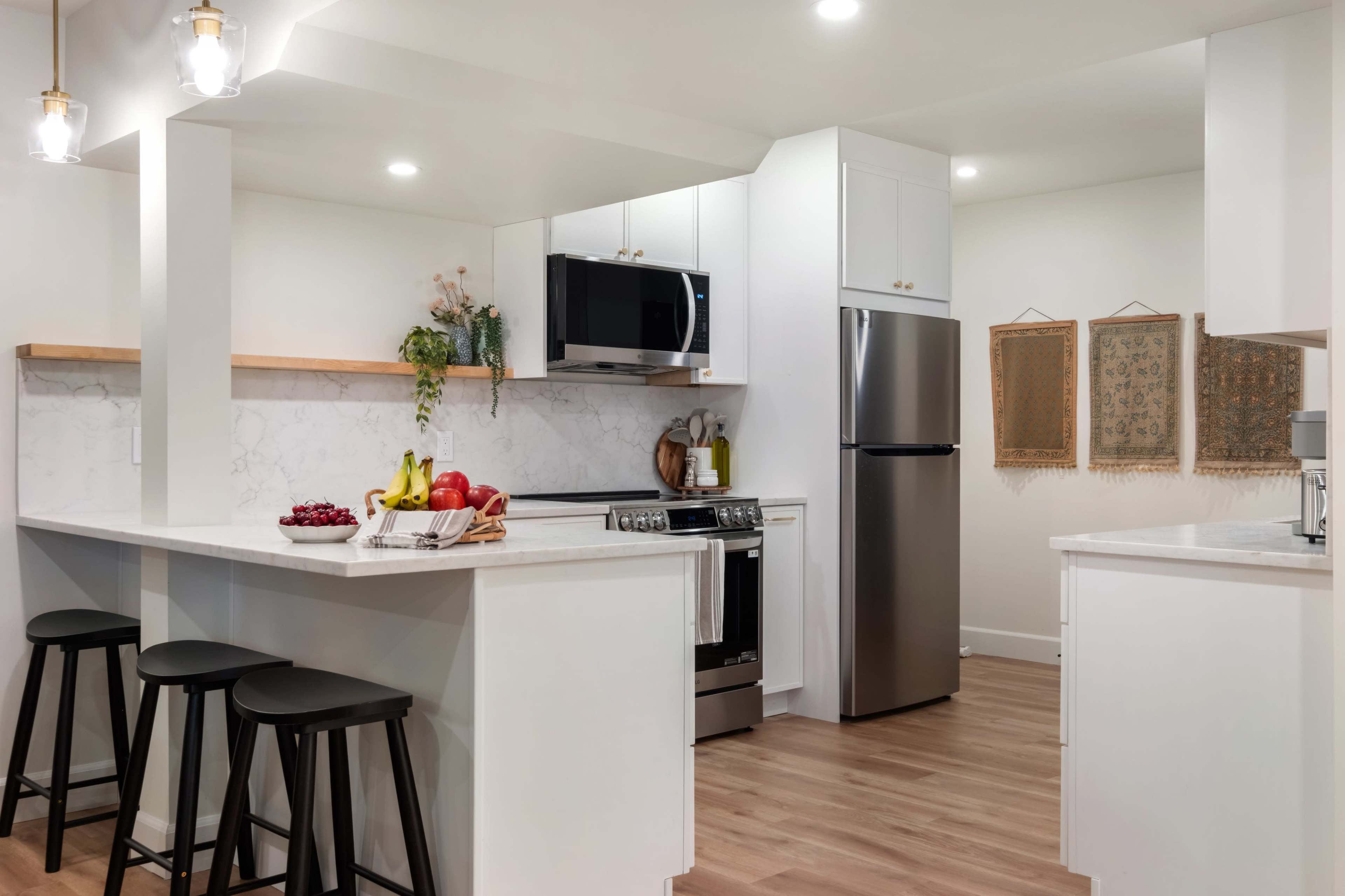 The image shows a modern kitchen with a central island, featuring black bar stools, stainless steel appliances, and decorative wall hangings.