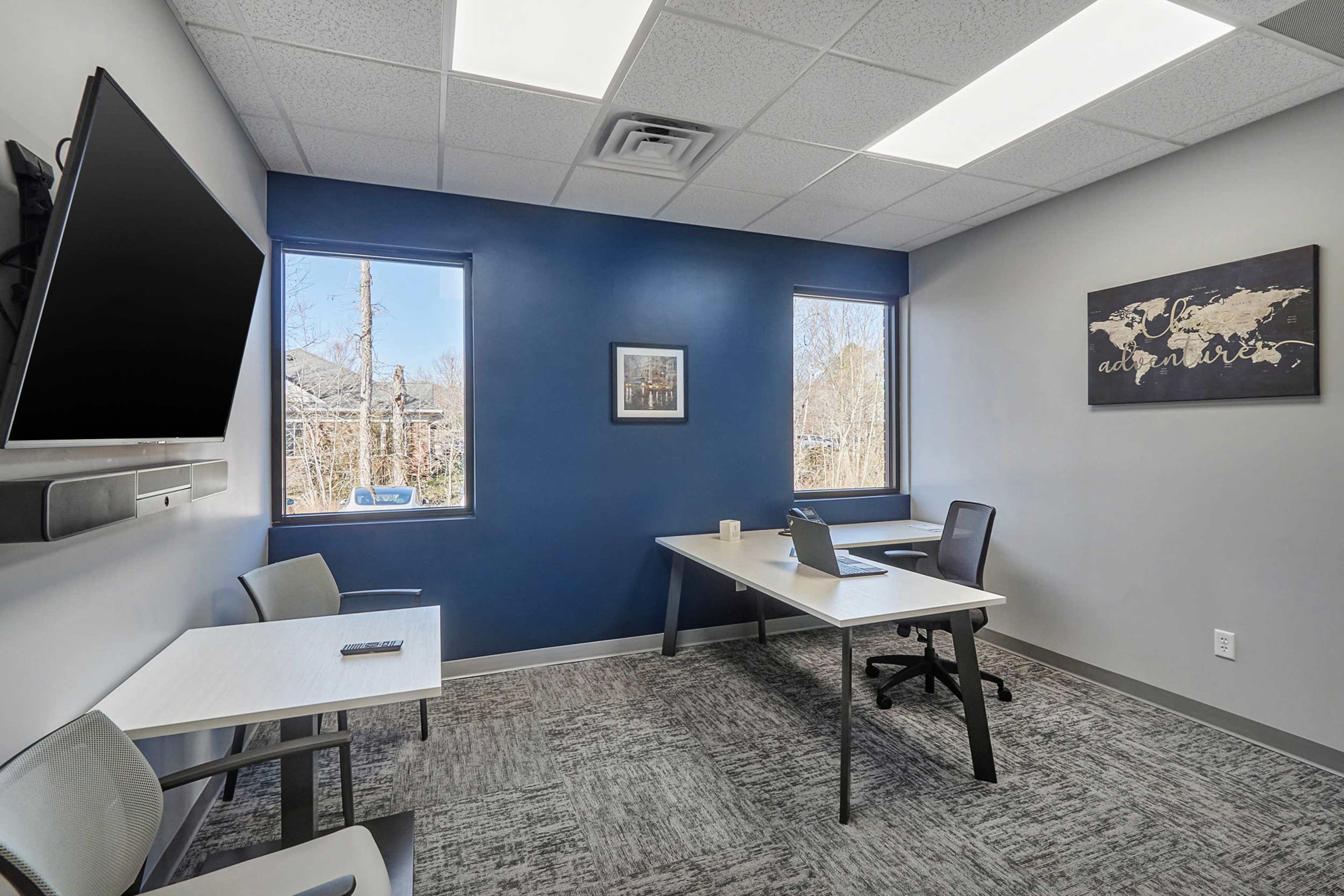 A modern office space with two windows, a desk with a laptop, and two chairs.