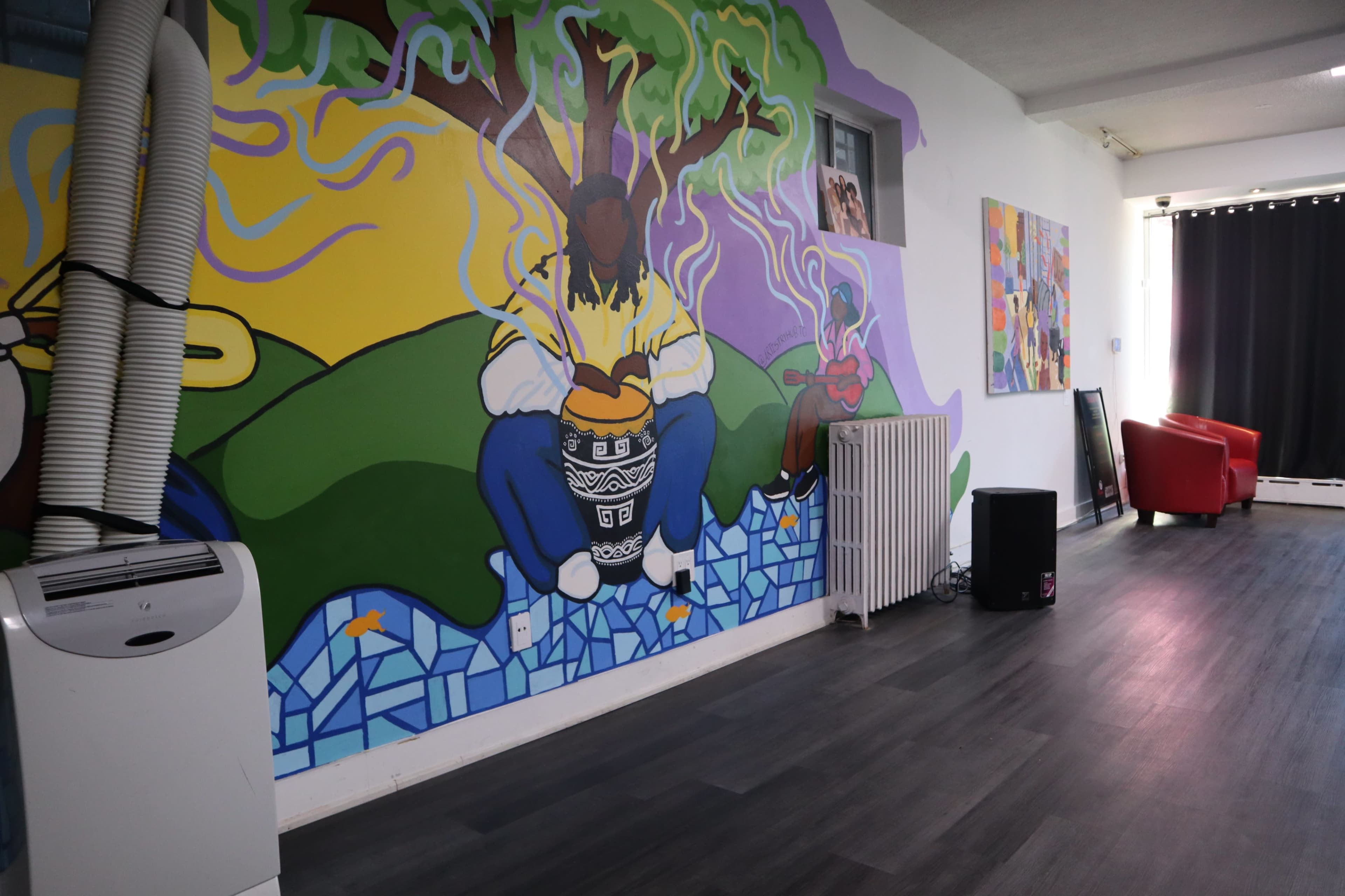 The image shows a brightly colored mural depicting a person playing an instrument beneath a tree, with a modern interior space featuring a red chair, a radiator, and an air conditioning unit.