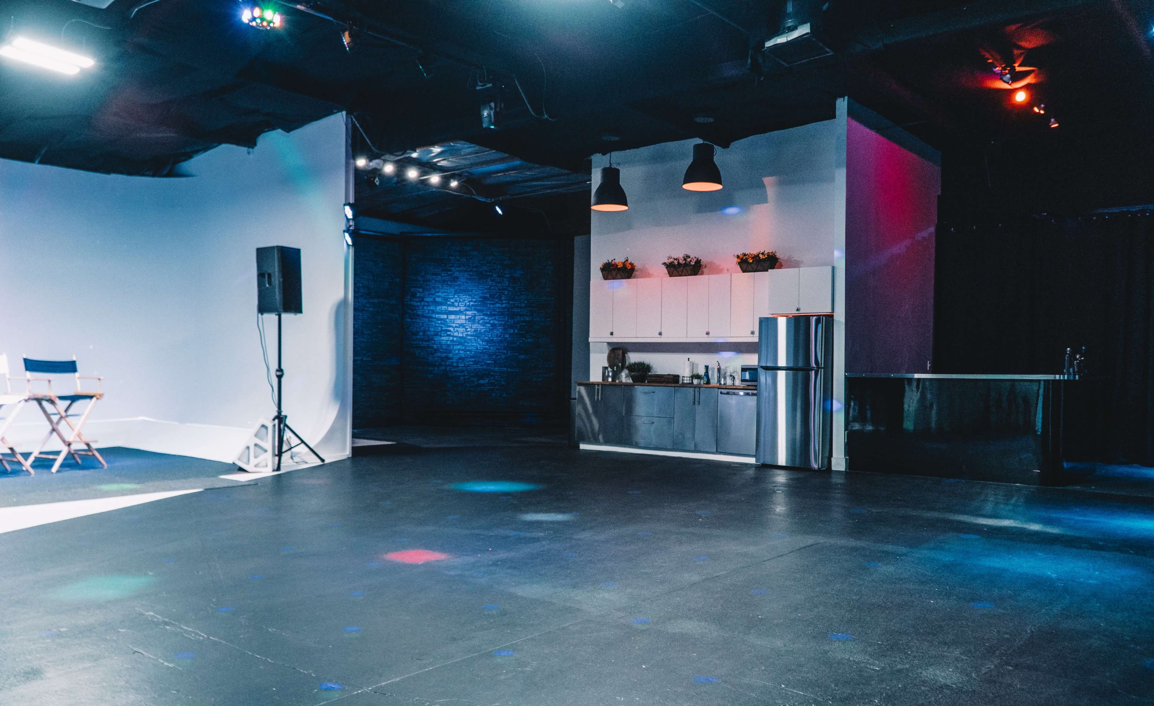 The image shows a spacious studio with a kitchen area on one side, featuring a refrigerator and cabinets, and a large empty floor area with colorful lights.