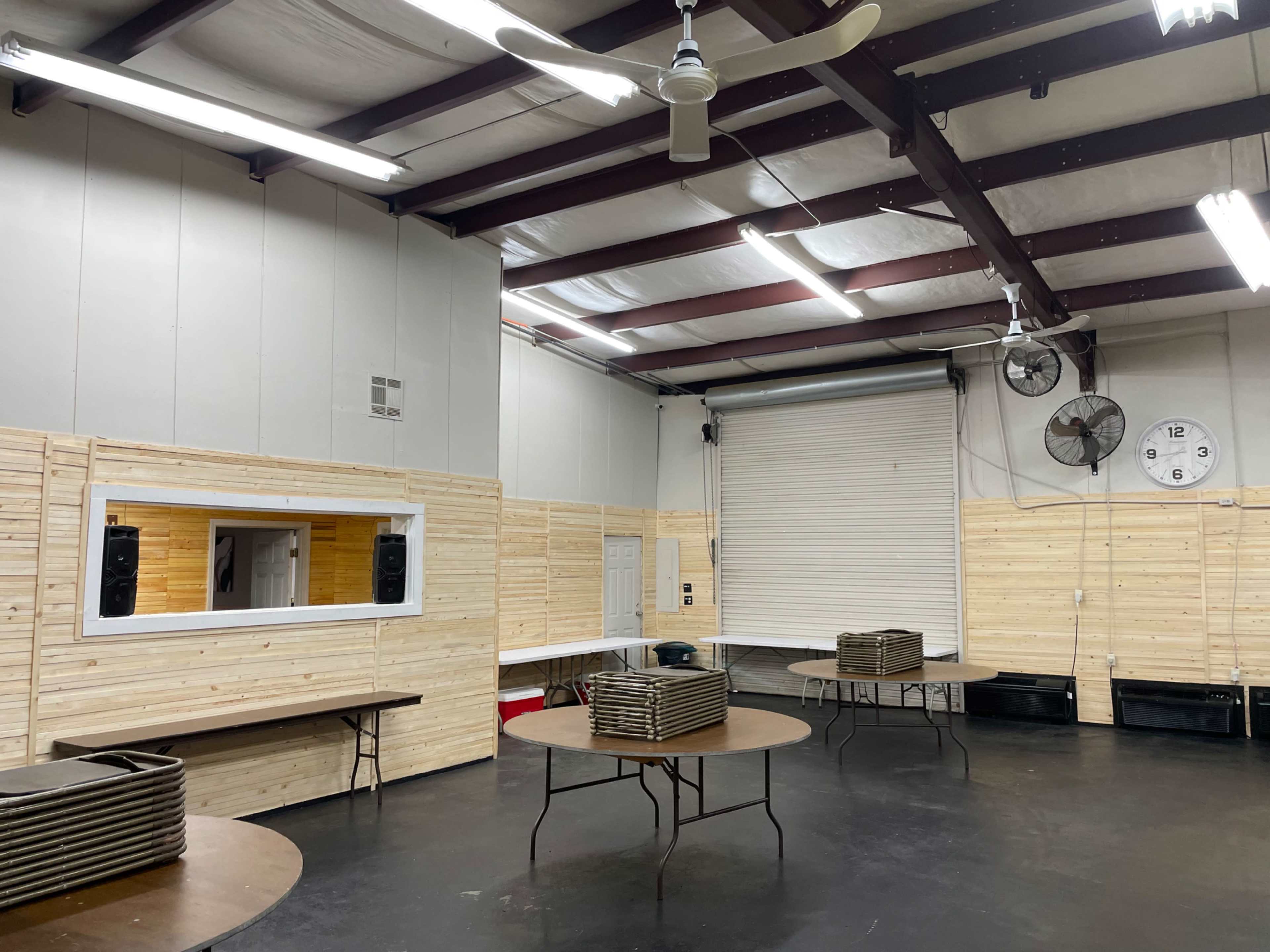 A spacious indoor area with wooden paneling, round tables stacked with trays, a large roll-up door, and wall-mounted fans.