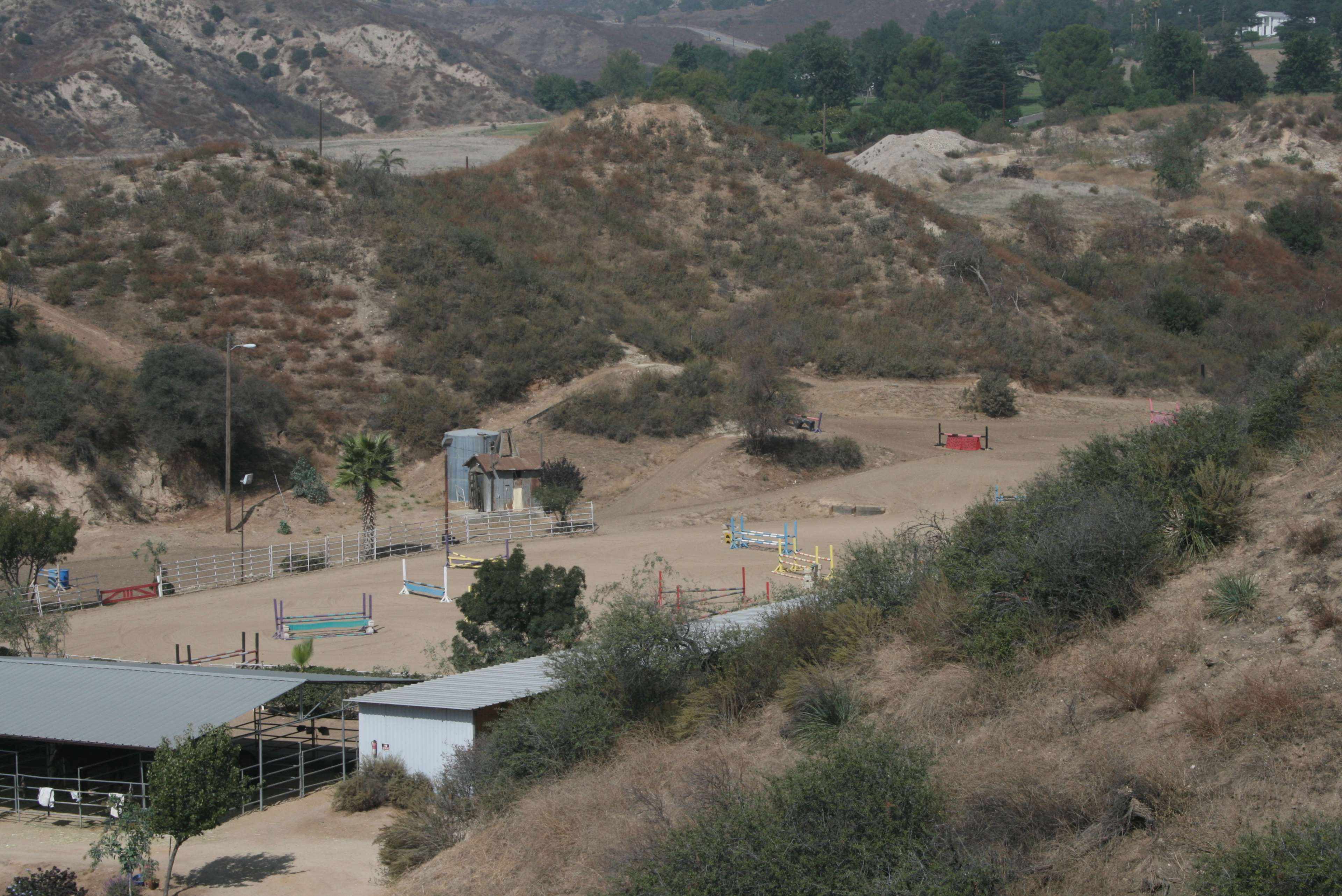 The image shows a horse riding arena situated in a dry, hilly landscape, surrounded by sparse vegetation and a few structures.