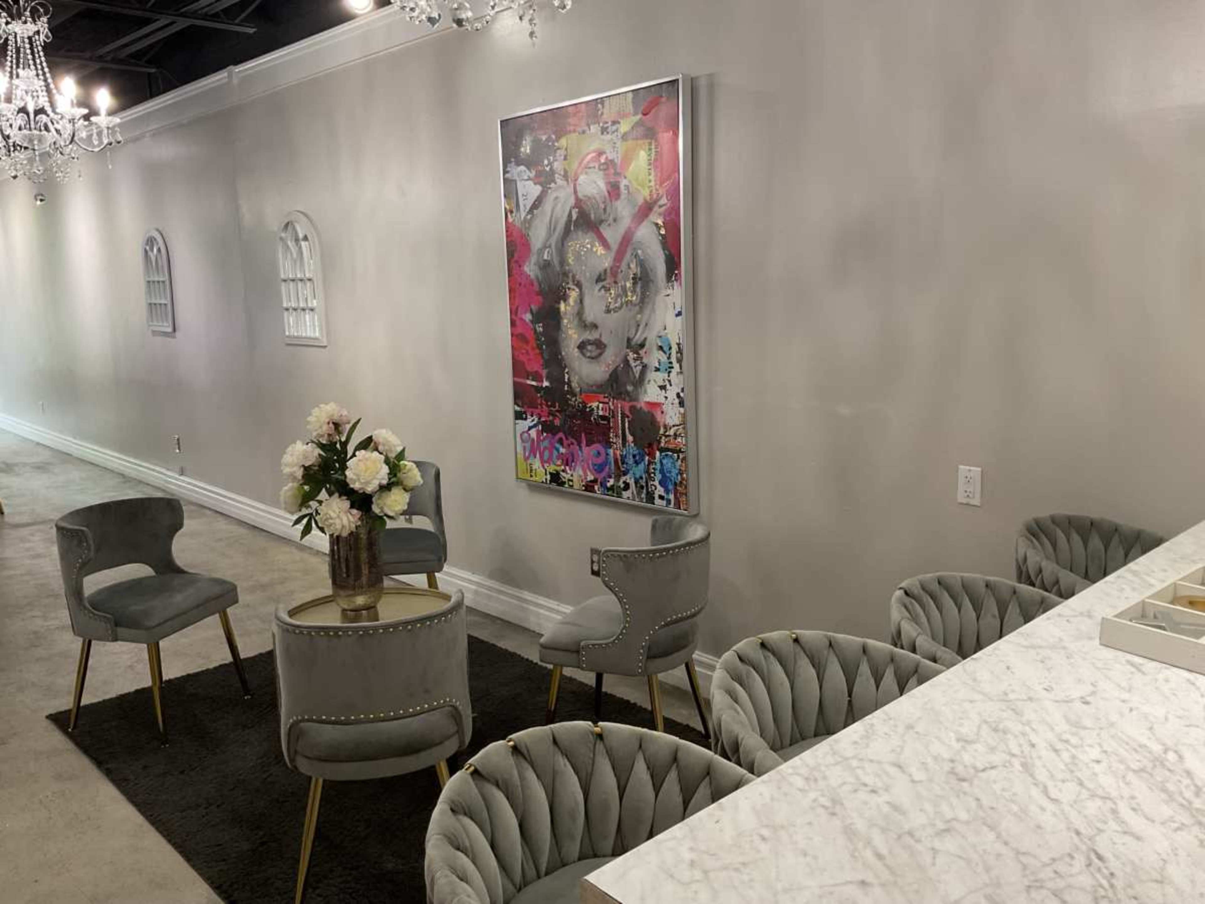 The image shows a modern interior with a gray color scheme, featuring a wall adorned with a colorful piece of art, several stylish chairs, a circular table with a floral arrangement, and a marble countertop.