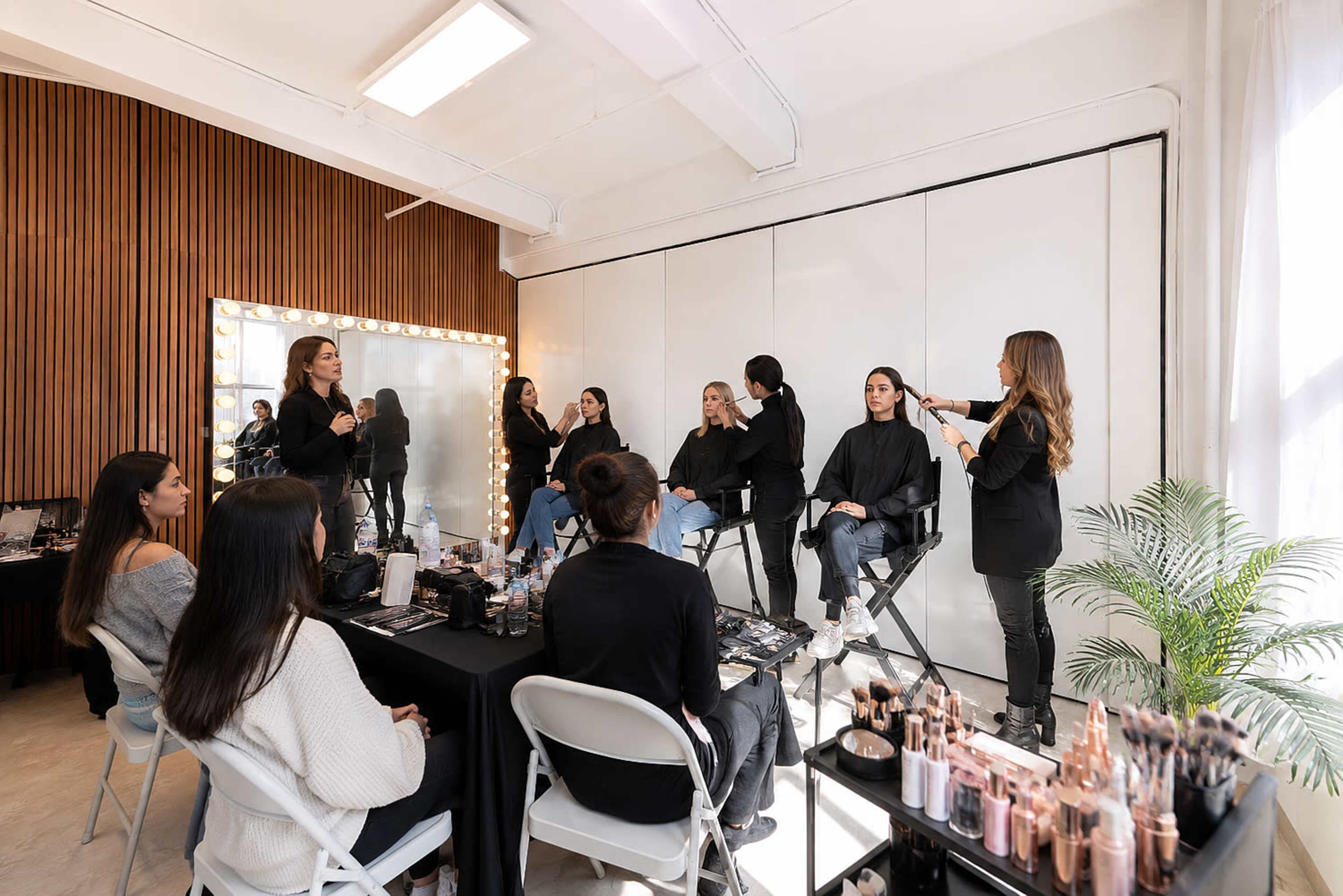 A group of makeup artists is providing makeup application to several clients in a well-lit studio with a large mirror and makeup displays.