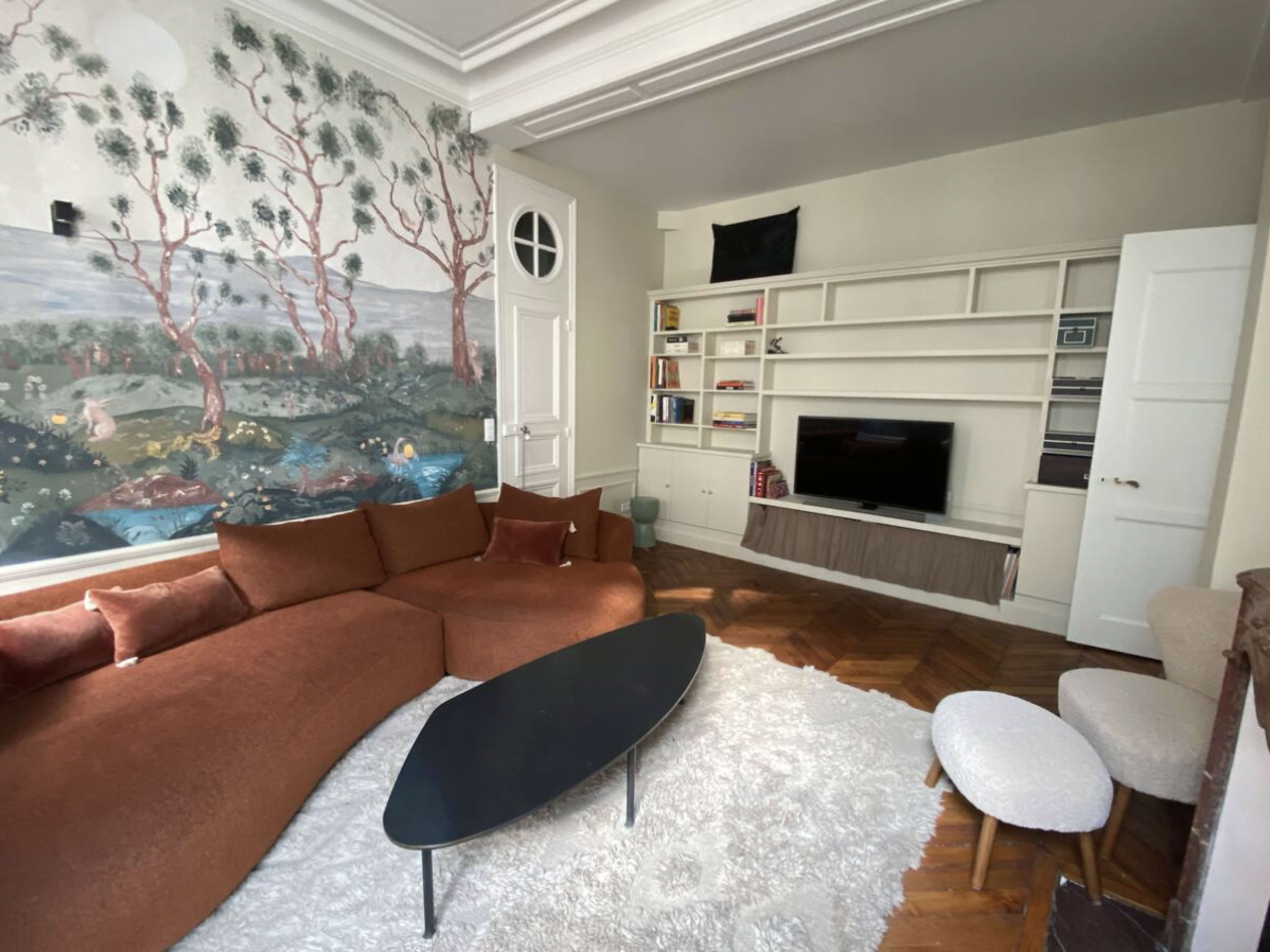 The living room features a curved brown sofa, a black coffee table, white fluffy rug, and a mural of trees and a landscape on one wall.