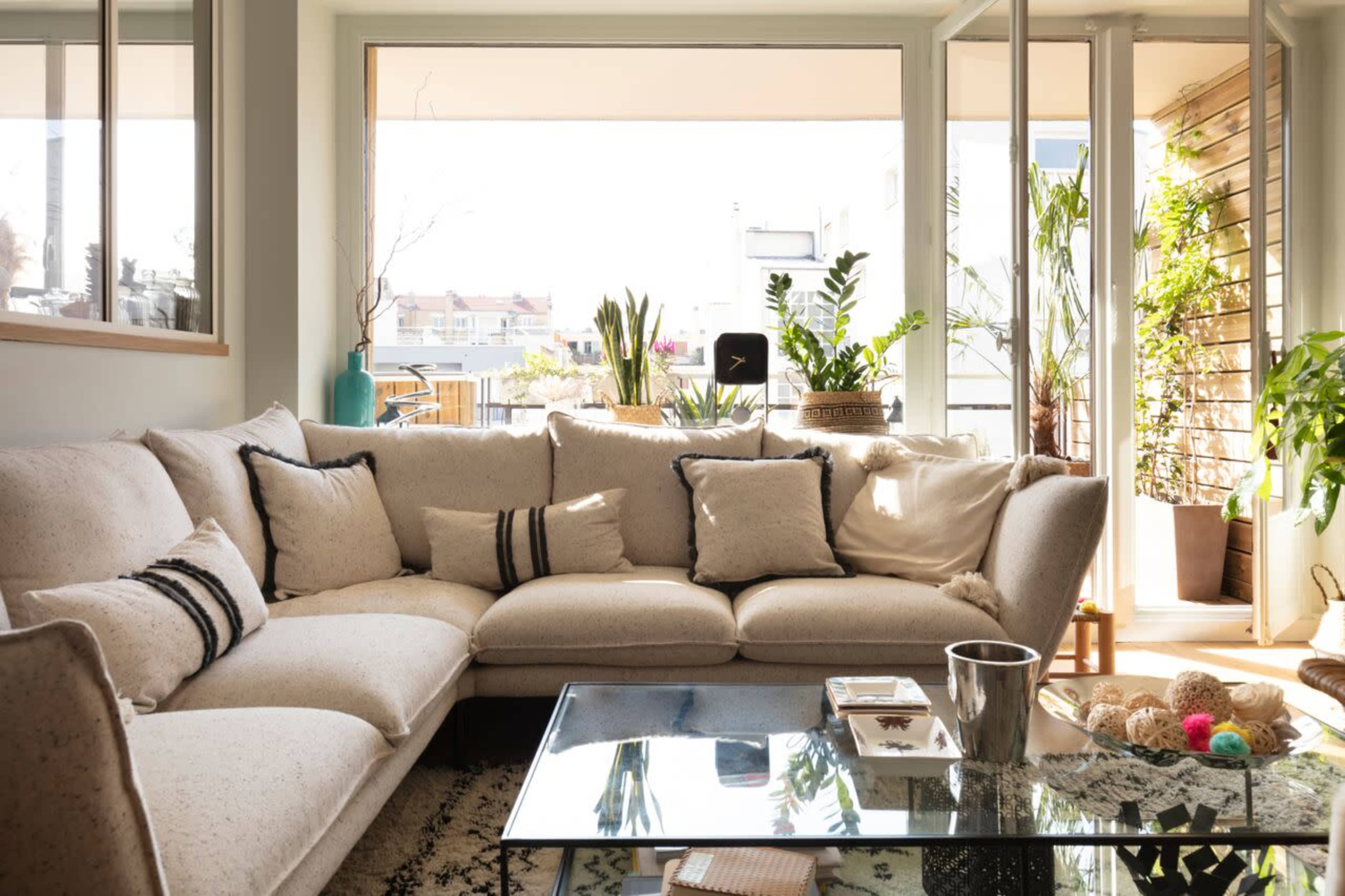 The image shows a bright living room with a large beige sectional sofa, a glass coffee table, and various indoor plants visible through the large windows.