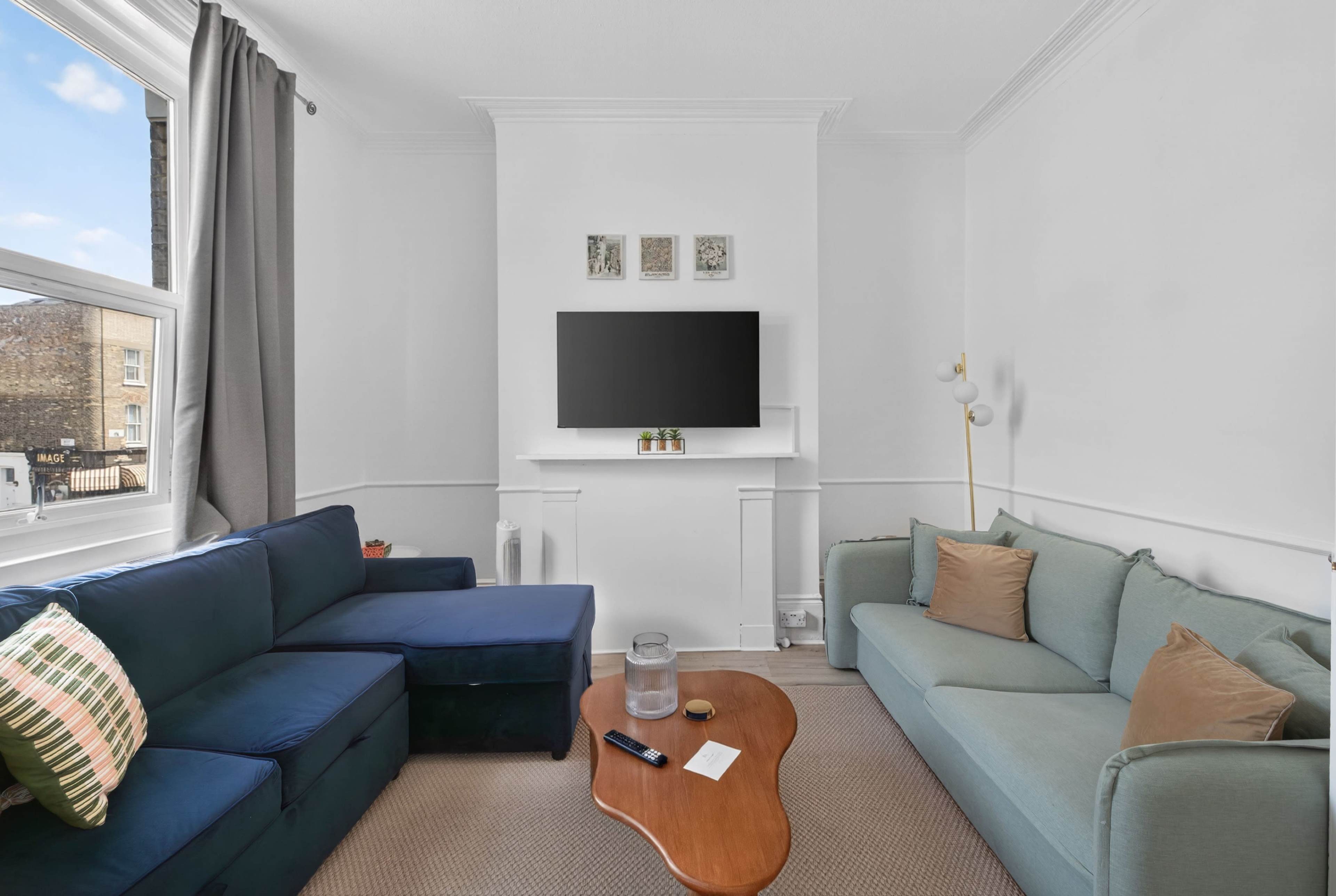 The image depicts a modern living room with a blue sectional sofa, a light green couch, a wooden coffee table, and a wall-mounted TV above a white mantel.